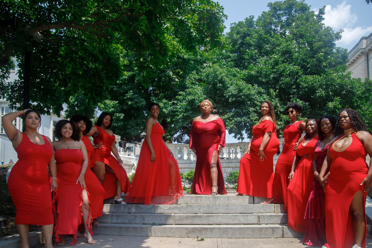 Group of women in coordinating red dresses pose on stone steps surrounded by lush green trees outdoors.
