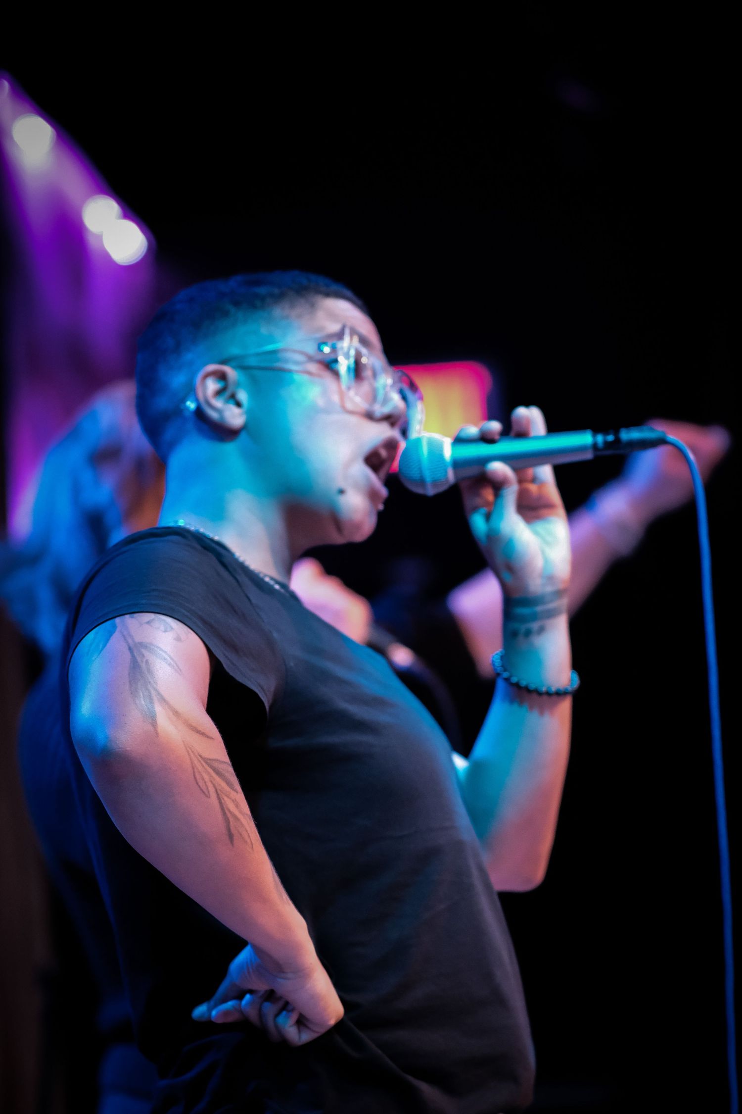 Male singer with glasses and tattoos performing on stage under blue stage lighting.