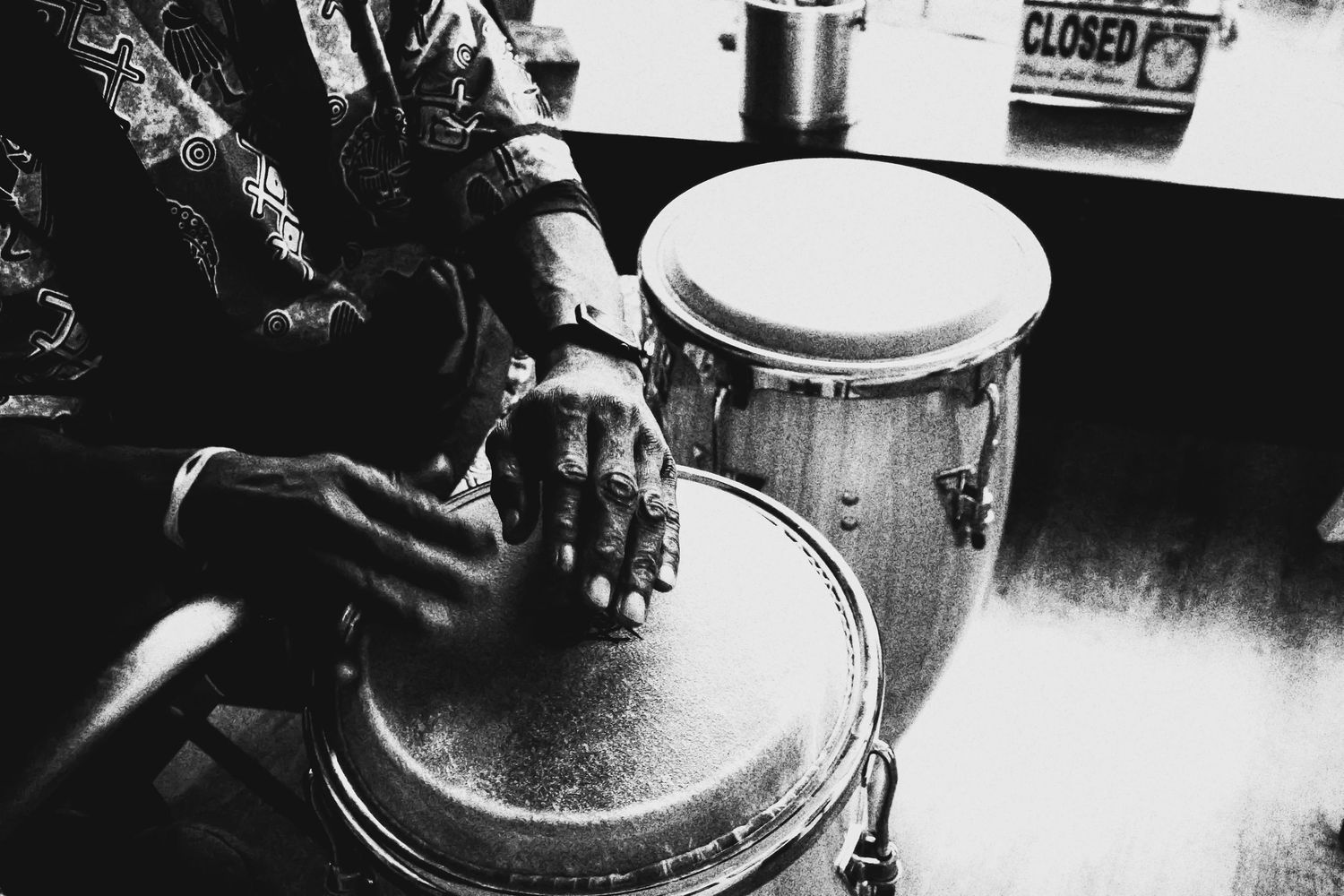 Black and white close-up of hands playing conga drums with a closed sign visible in background.