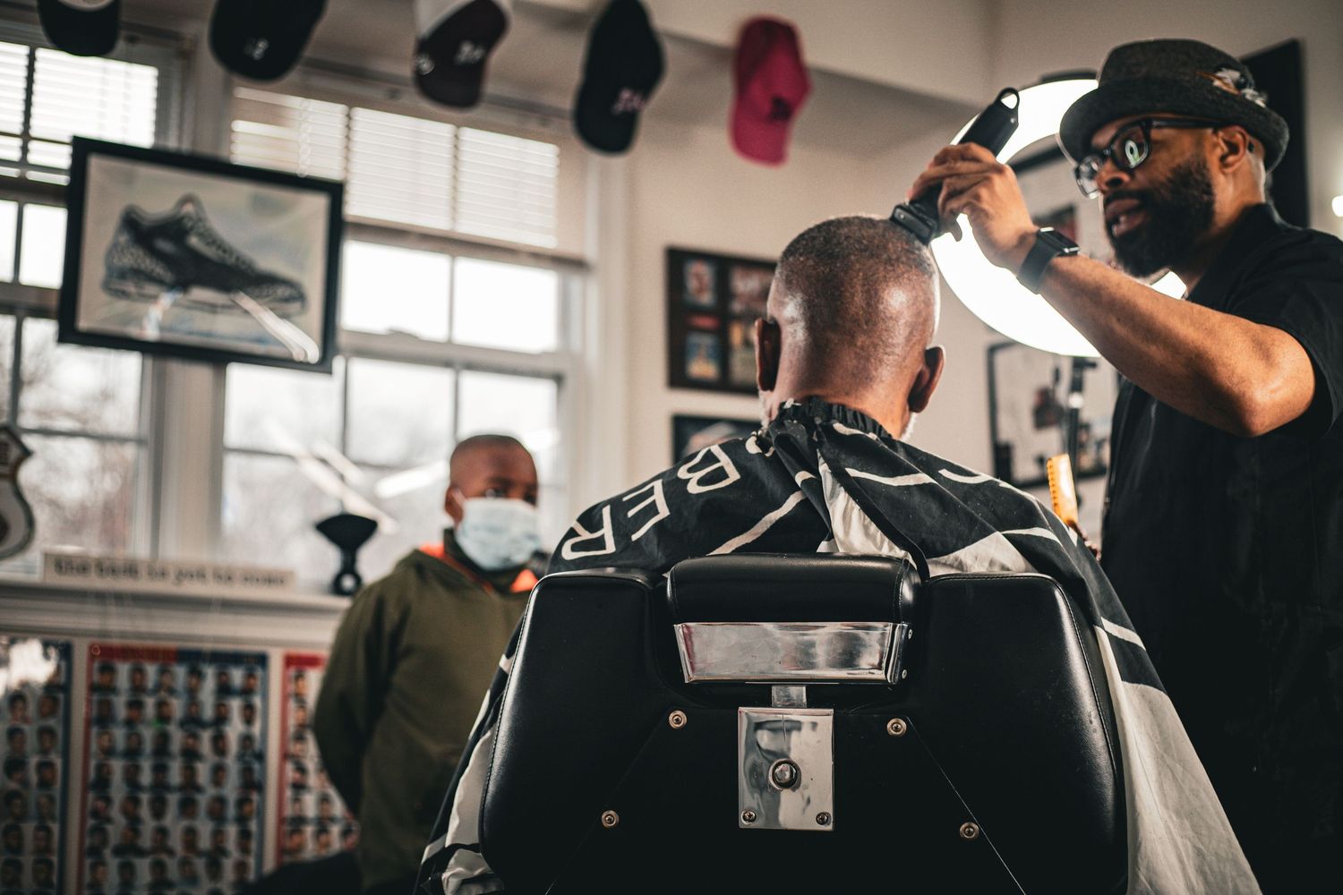 A barber styles a client's hair in a busy barbershop while a masked observer watches nearby.