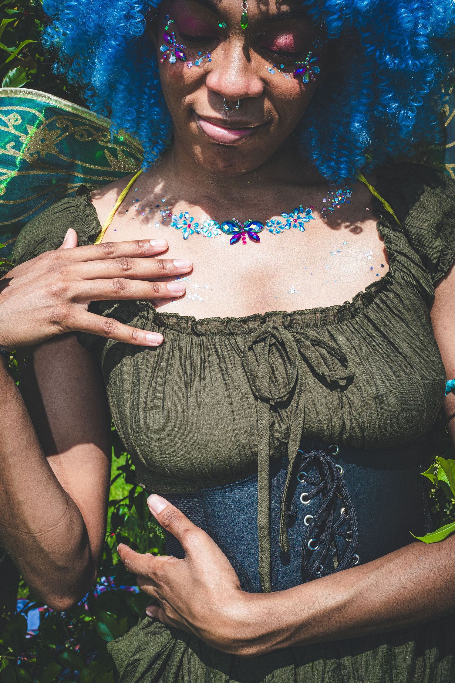 Woman in green corset top with blue gem body art and blue wig poses in fantasy fairy costume.