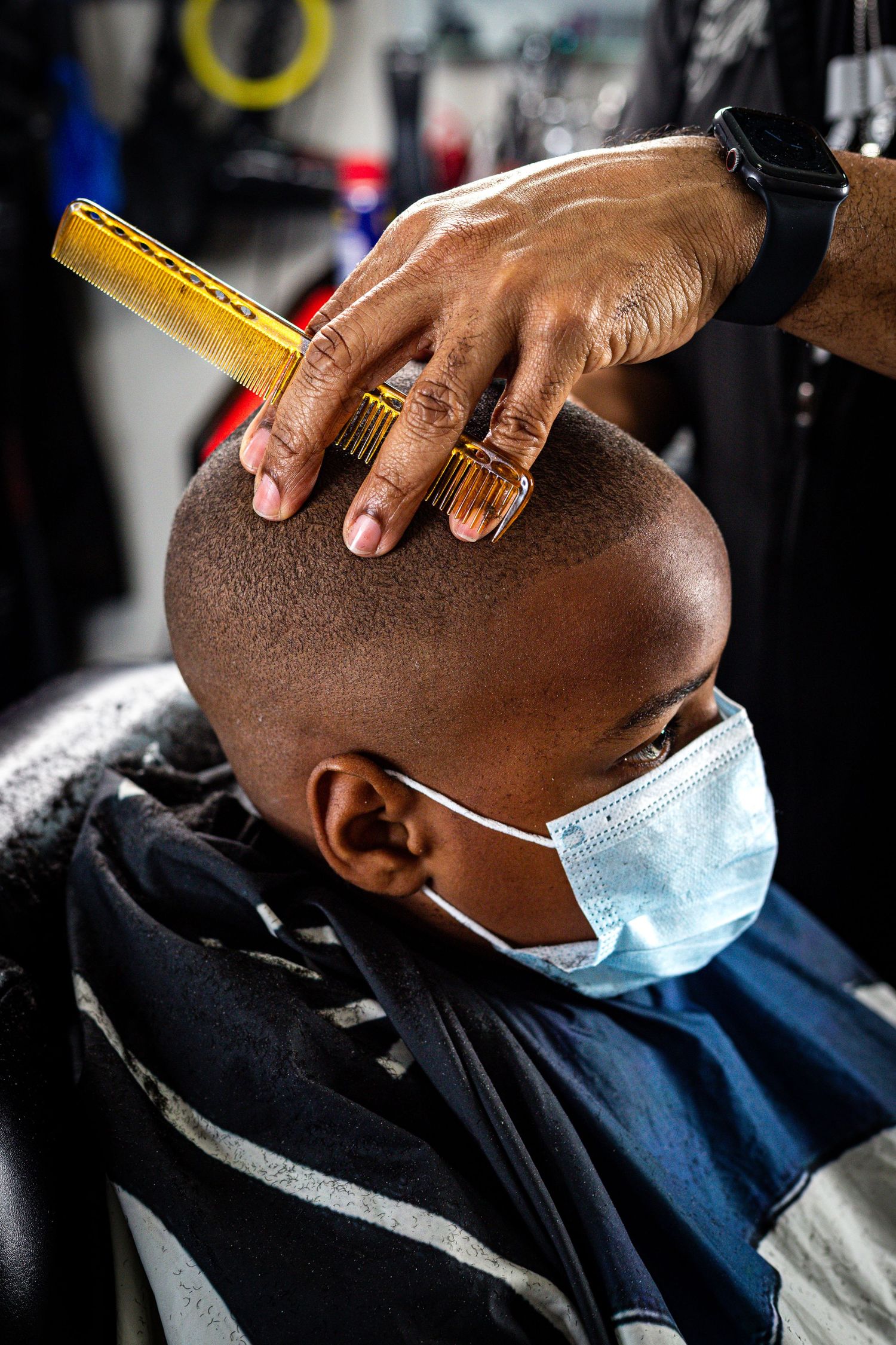 Barber using a comb on a young boy wearing a face mask during a haircut.