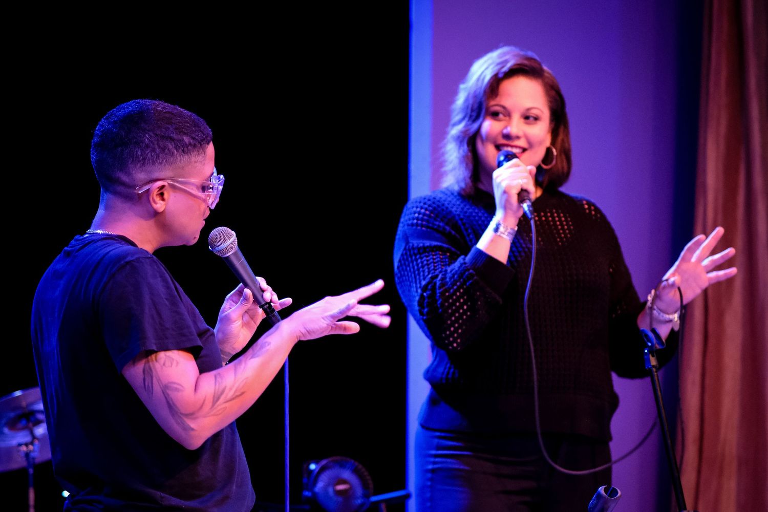 Two performers singing on stage with microphones under purple spotlight lighting.