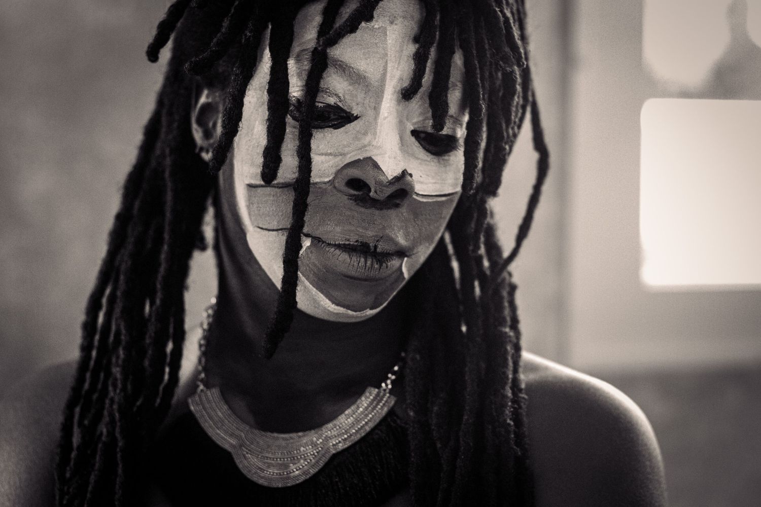 Black and white portrait of person with dreadlocks and white face paint looking downward.
