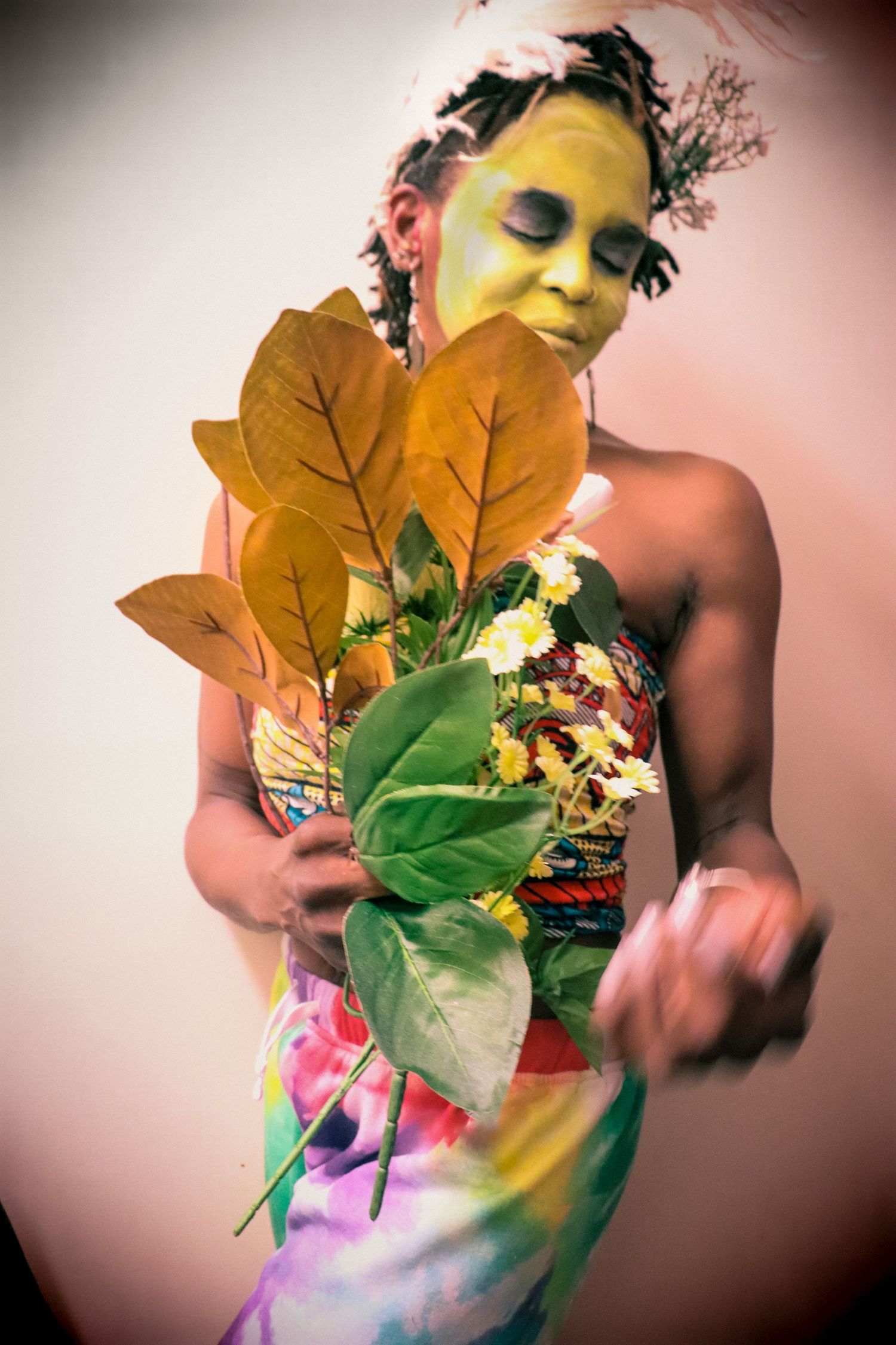 Woman with yellow face paint holding large tropical leaves and flowers in a colorful wrapped outfit.
