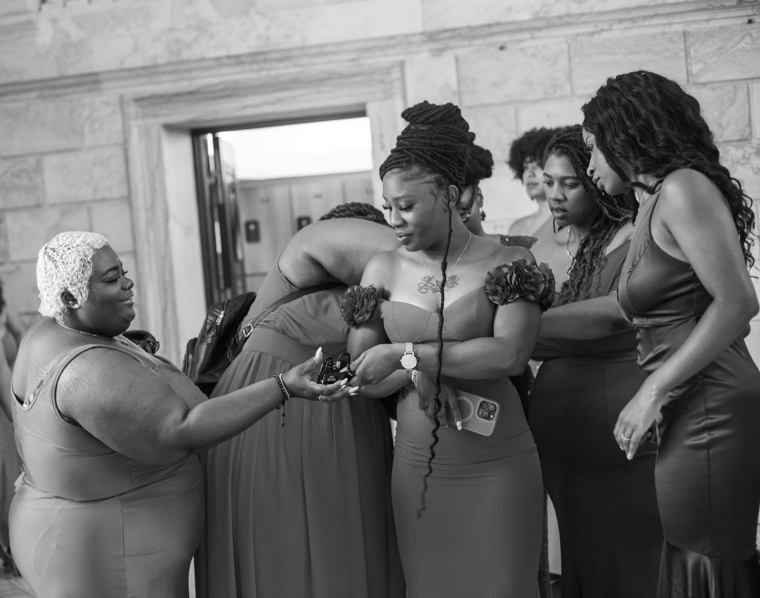 Black women who look like Bridesmaids in gray gowns gathered together examining jewelry in elegant marble building interior.