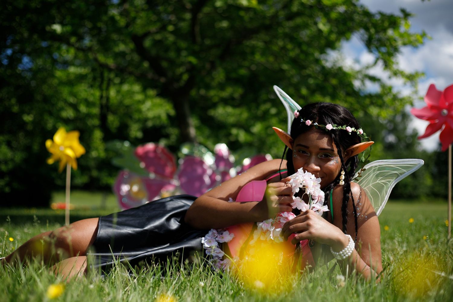 Young woman in fairy costume with wings lies in grass smelling white flowers at outdoor festival.