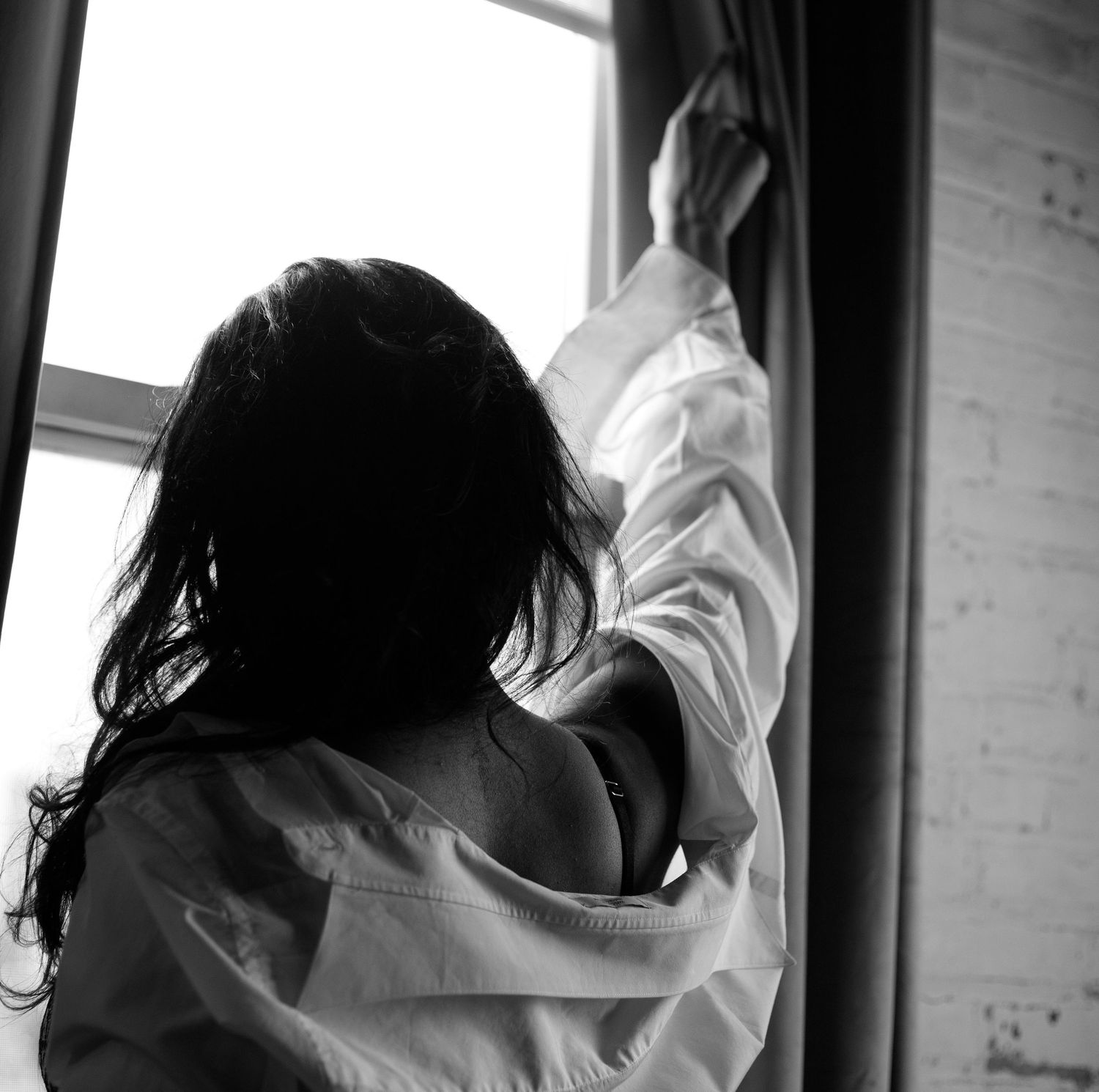 Silhouetted black and white photo of a woman in a white shirt reaching toward a bright window with curtains.