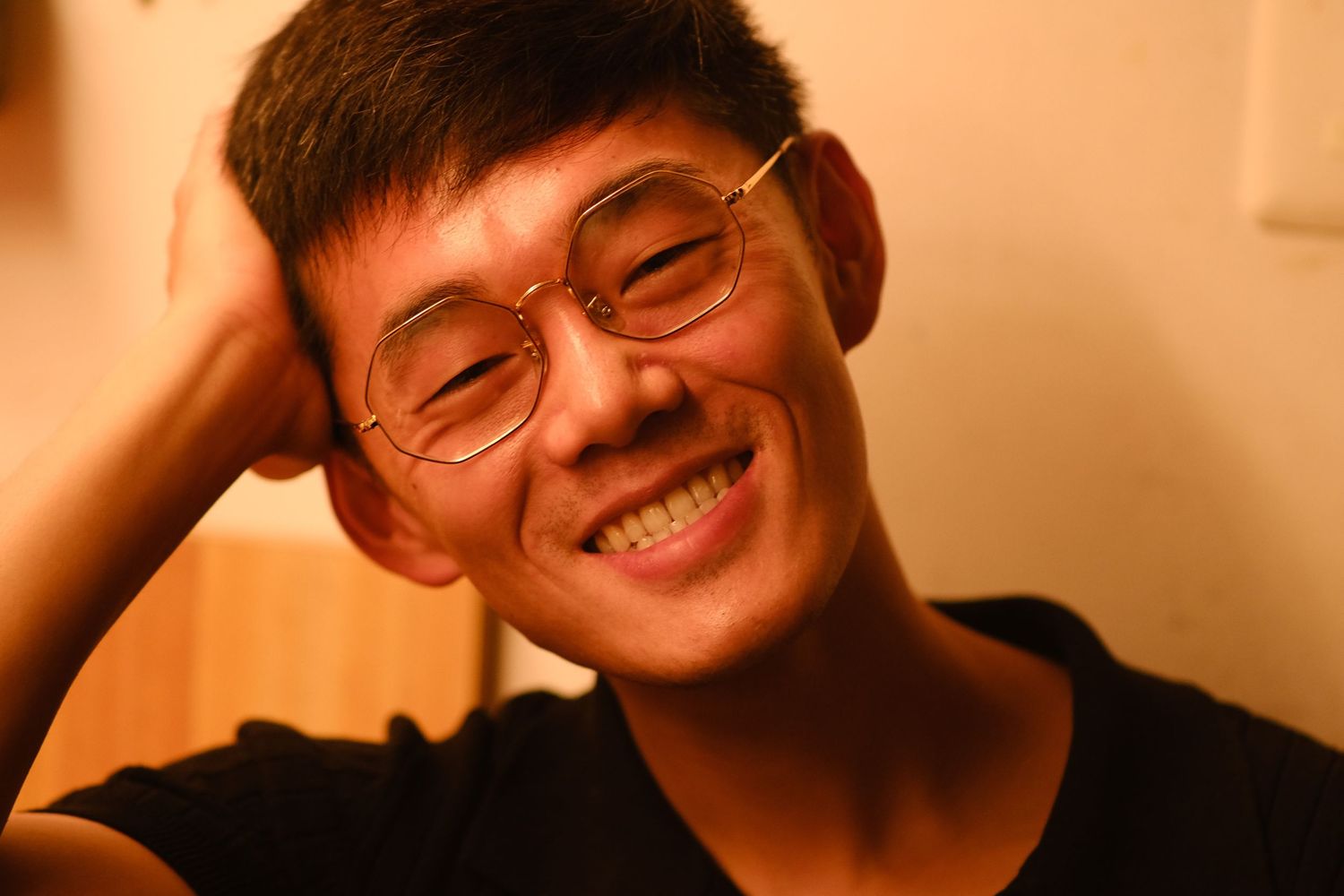 Smiling young Asian man wearing round glasses with hand behind head in warm amber lighting.