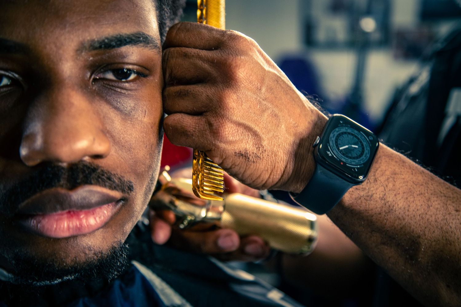 Close-up of a barber using gold clippers to cut a young mans hair with an Apple Watch visible.