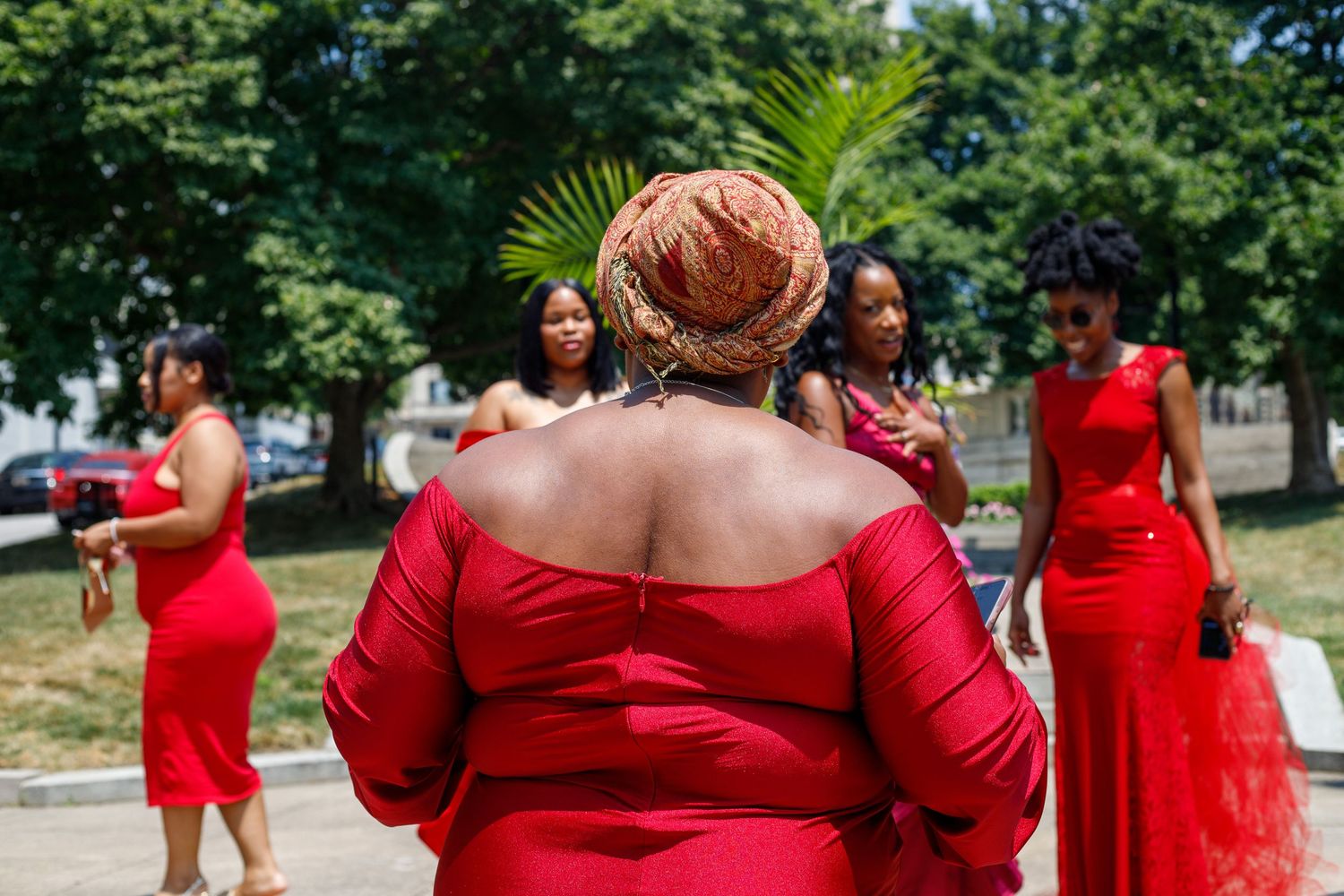 Group of women in matching red dresses gather outdoors on a sunny day with green trees in the background.