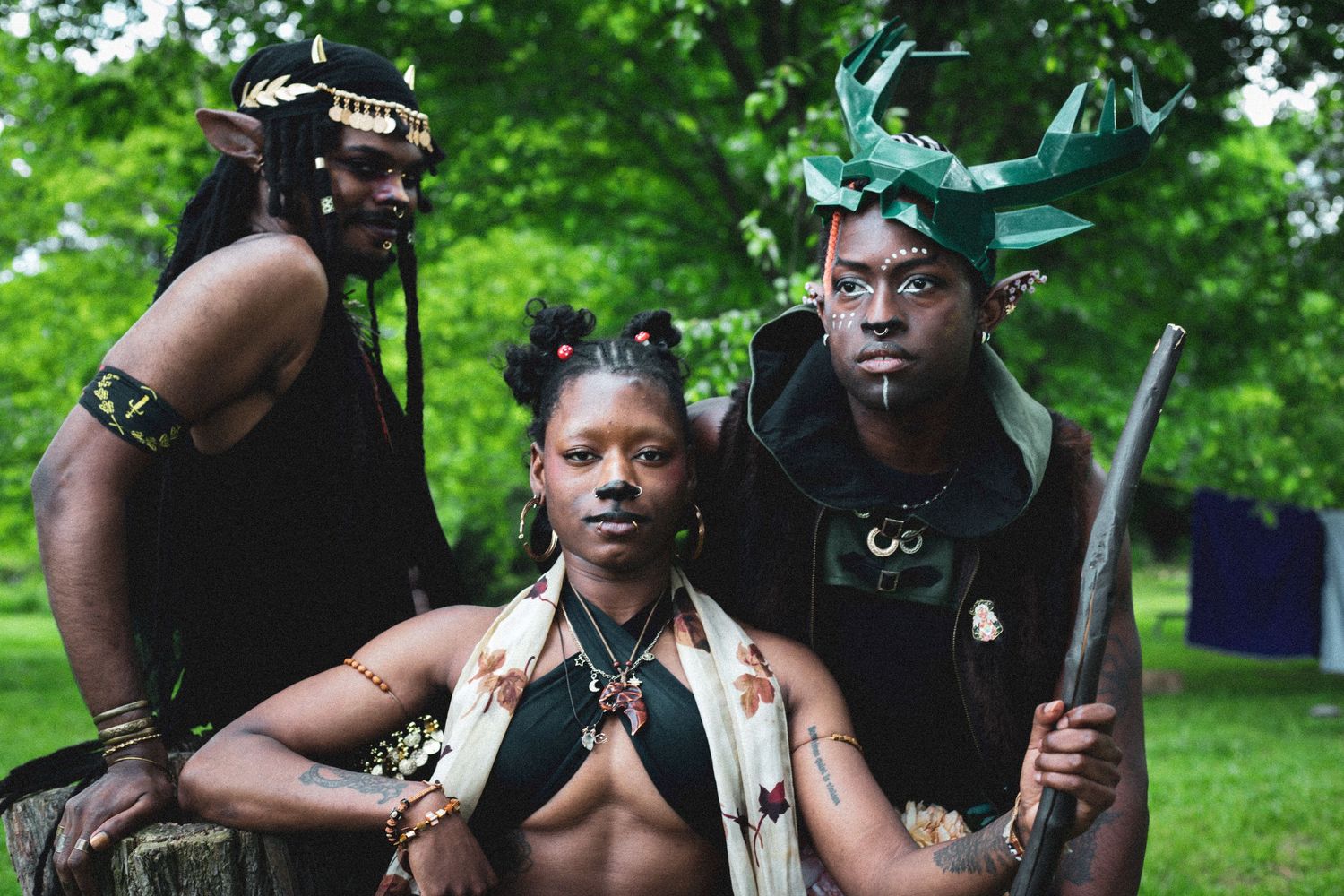 Three people in elaborate fantasy elf warrior costumes with face paint pose outdoors in park.