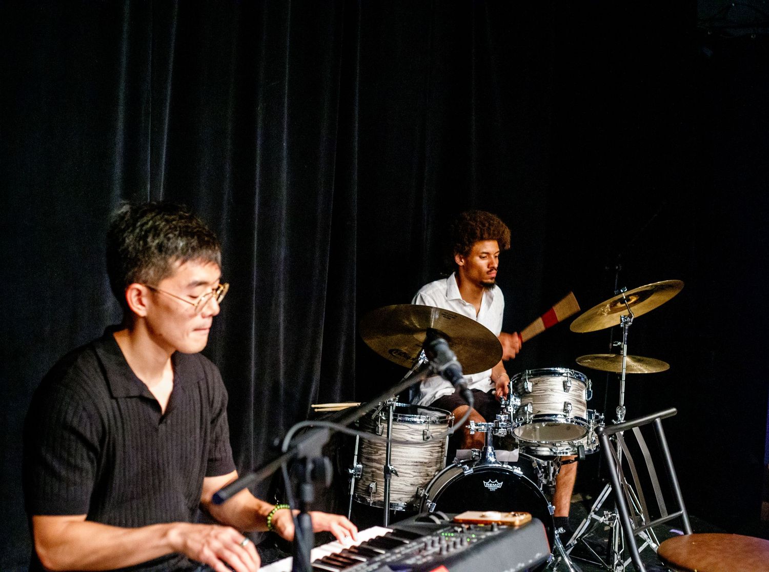 Two musicians performing live, one playing keyboard and another on drums against a dark curtain backdrop.