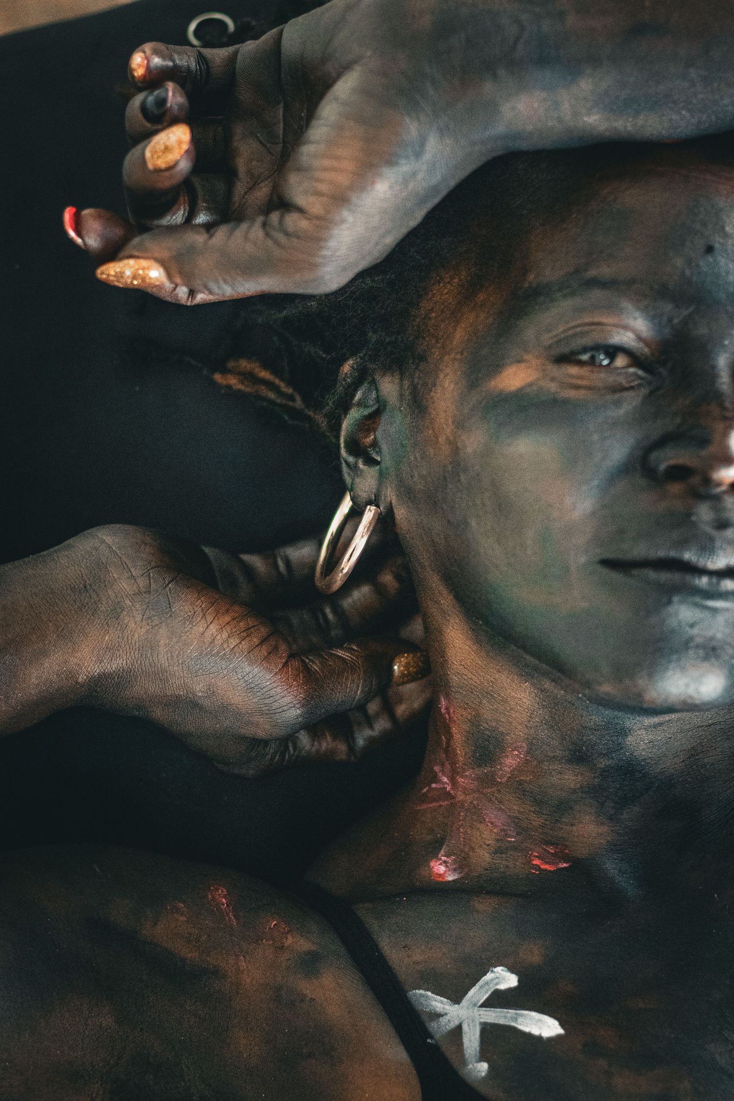 Close-up portrait of a model covered in dark body paint with hoop earrings and painted X symbol on chest.