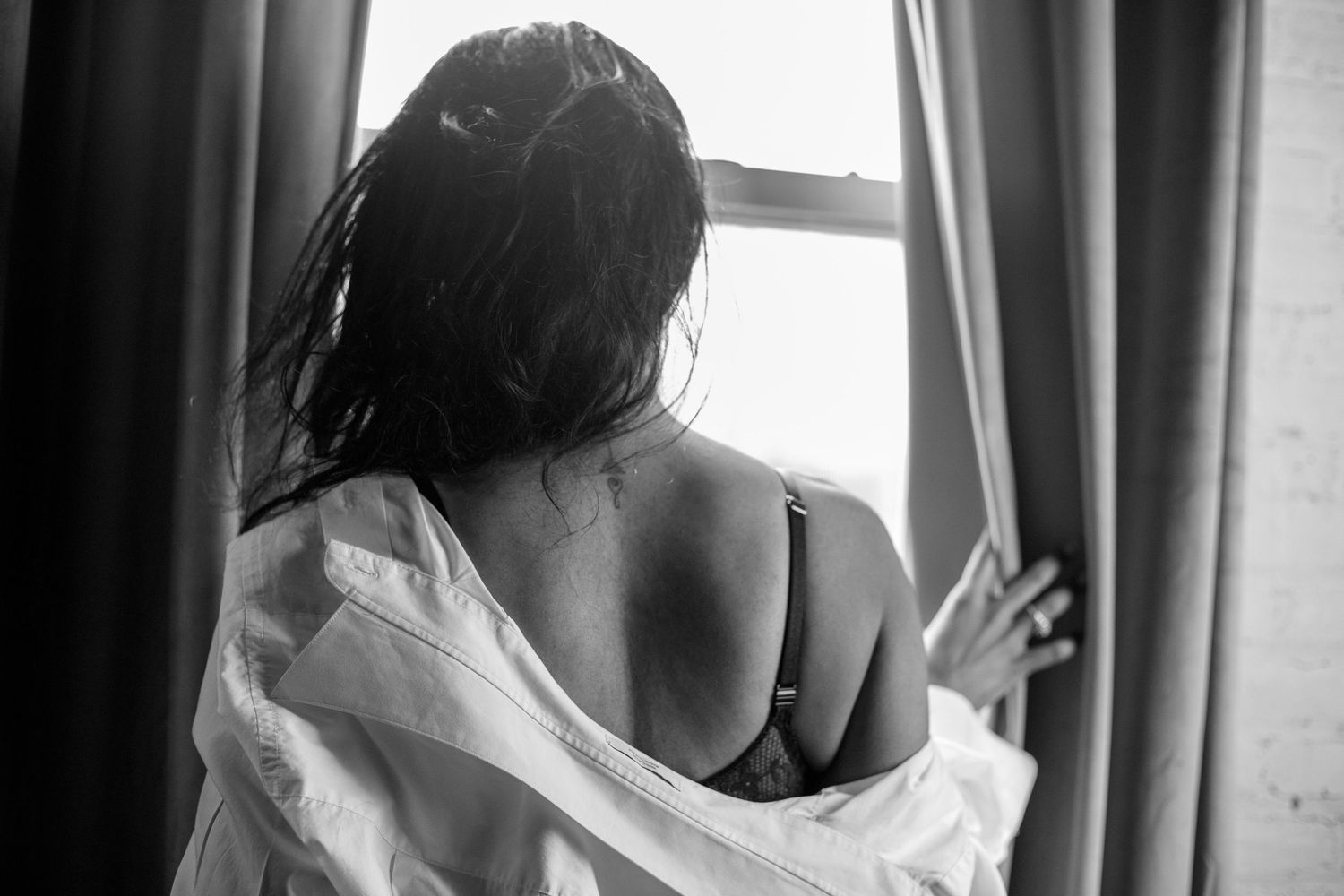Black and white boudoir image of a woman from behind in an open white shirt near a curtained window.