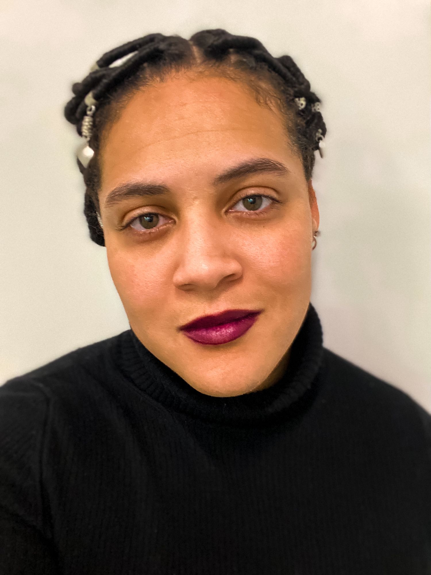 Close-up portrait of a woman with braided hair adorned with beads wearing a black turtleneck and dark lipstick.