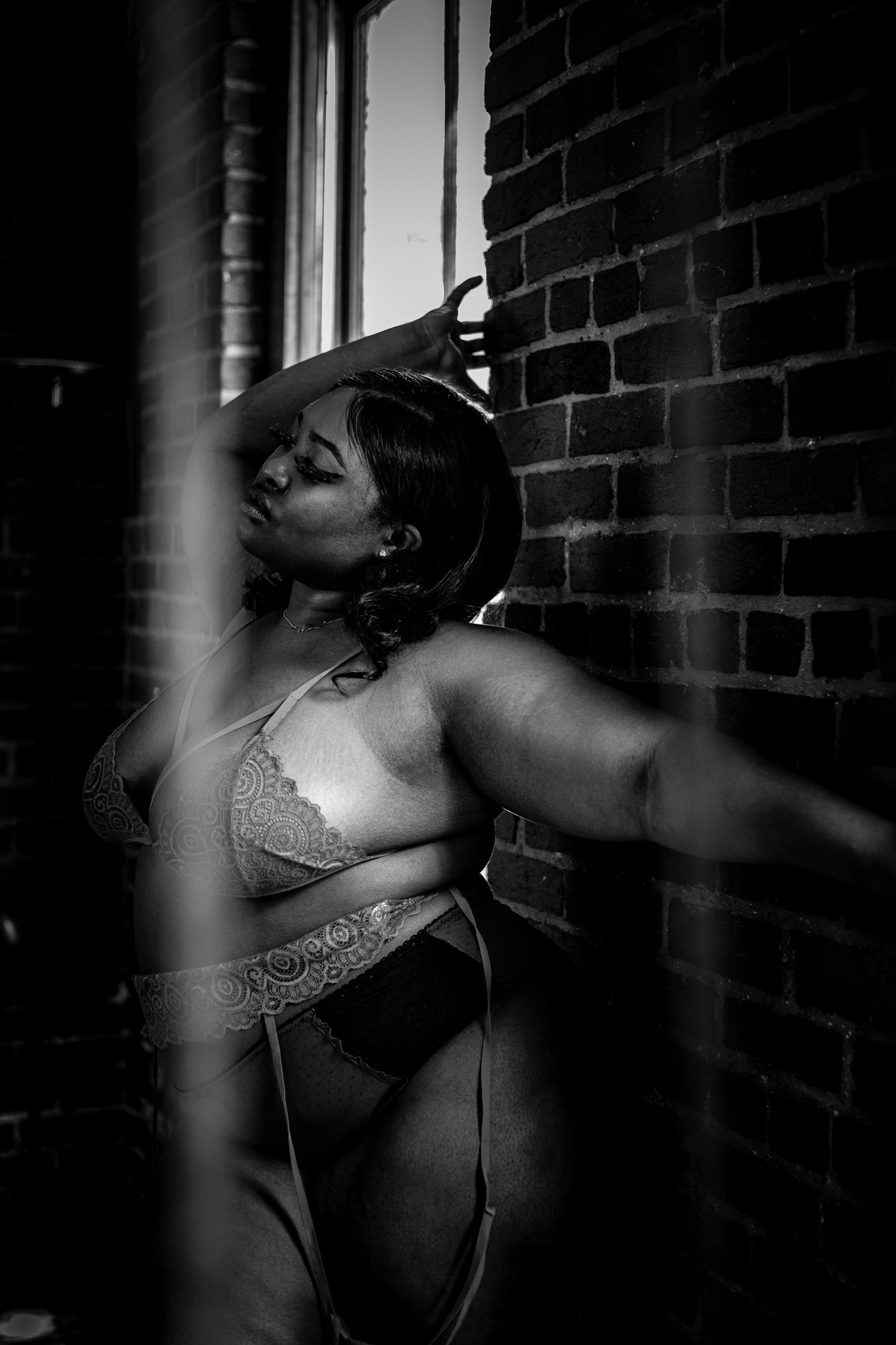Black and white boudoir portrait of a curvy woman in lingerie leaning against a brick wall by a window.