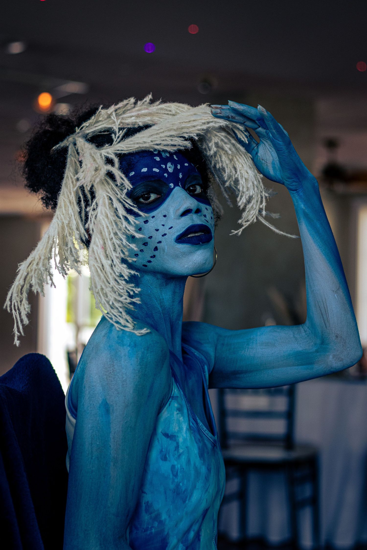 Model covered in blue body paint wearing white dreadlock wig and blue spotted mask at an event.