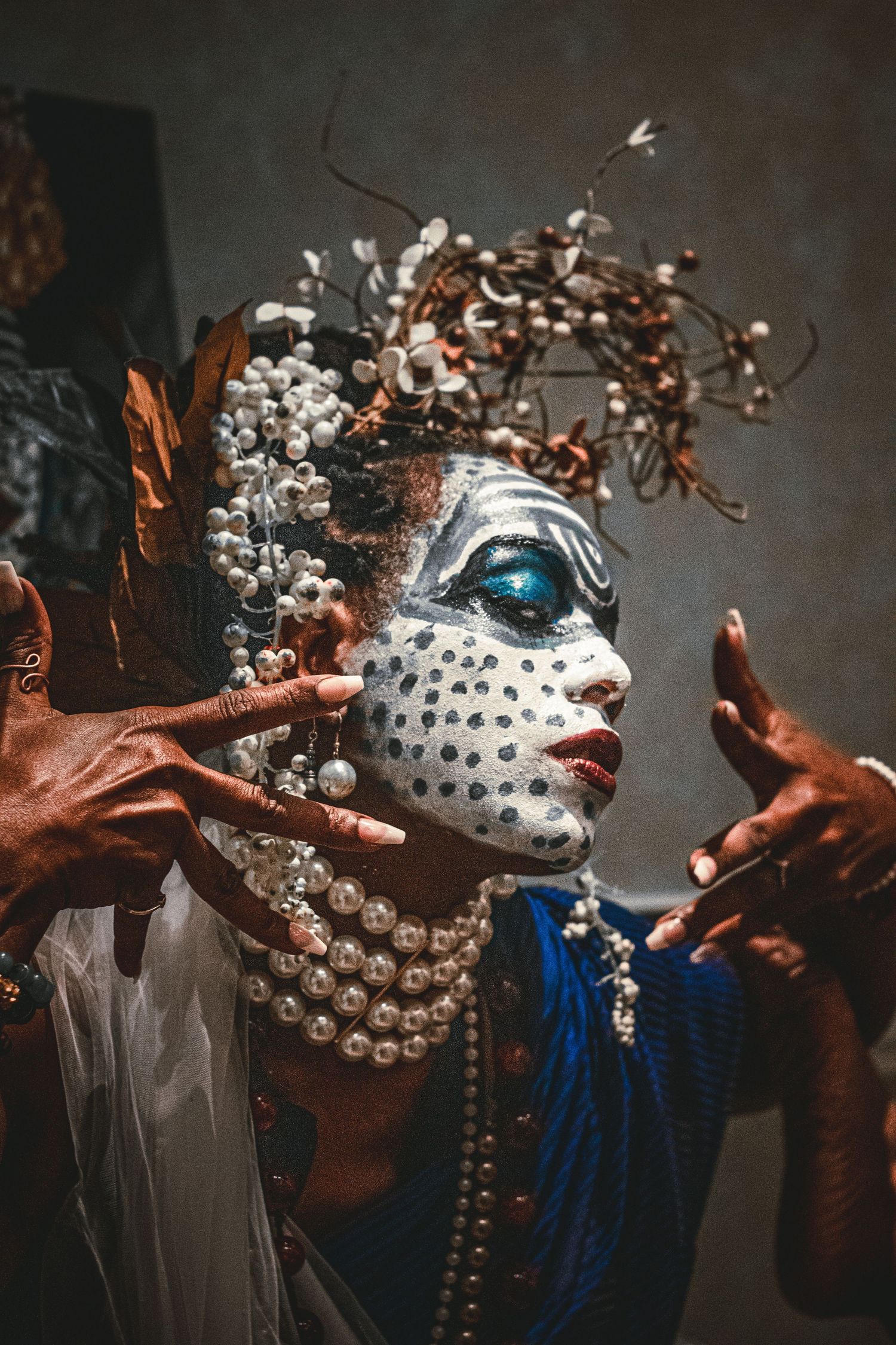 Person with elaborate white face paint, pearl necklaces, and ornate headdress of branches and beads poses dramatically.