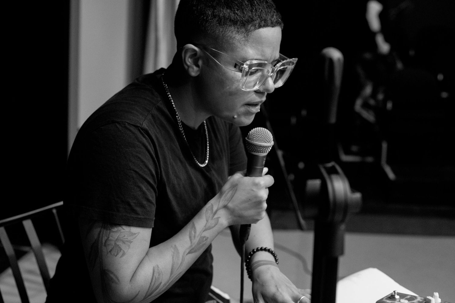 Black and white portrait of a man with glasses and tattoos speaking into a microphone at a music event.