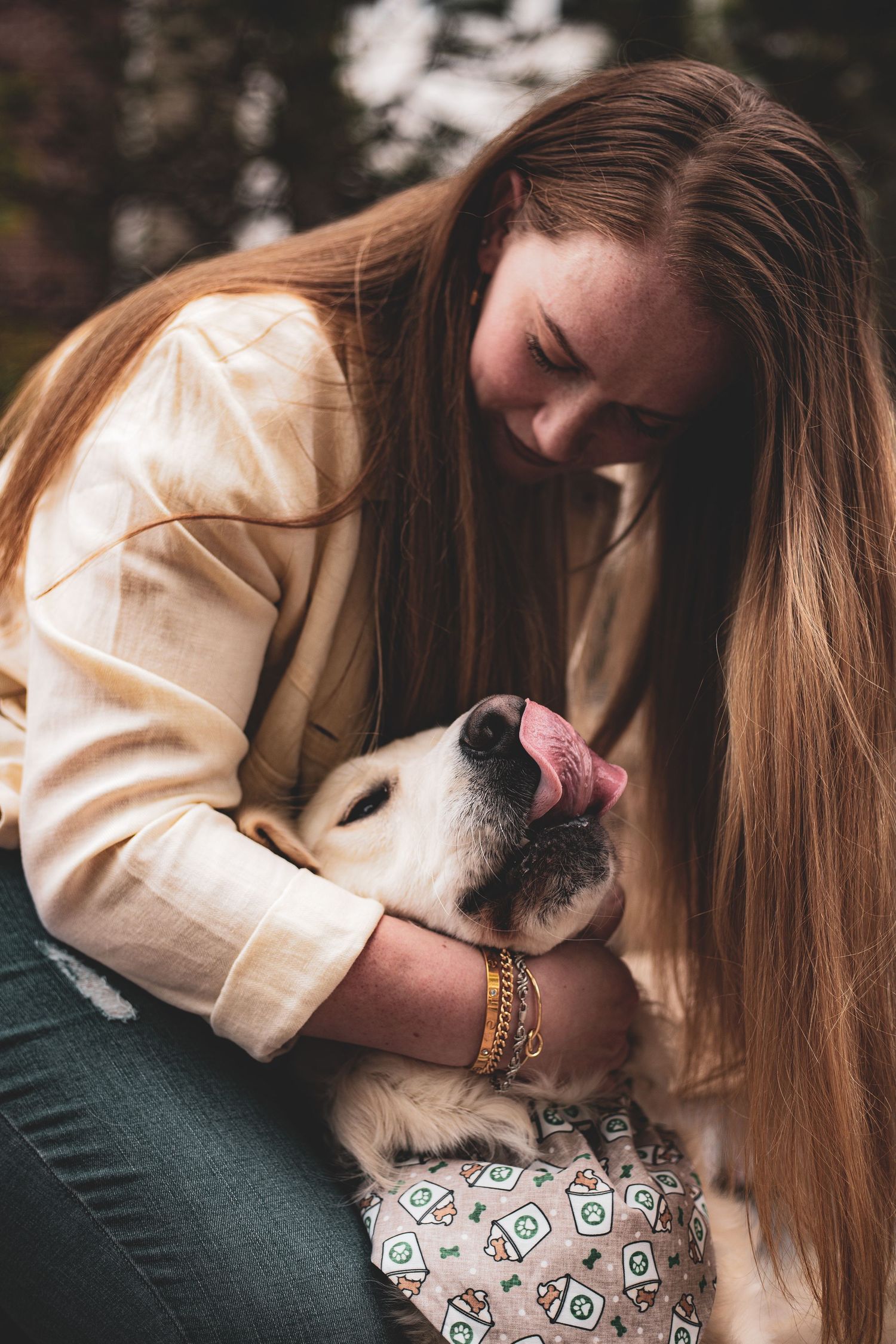 Long-haired woman in yellow jacket hugging a golden retriever licking her face outdoors.