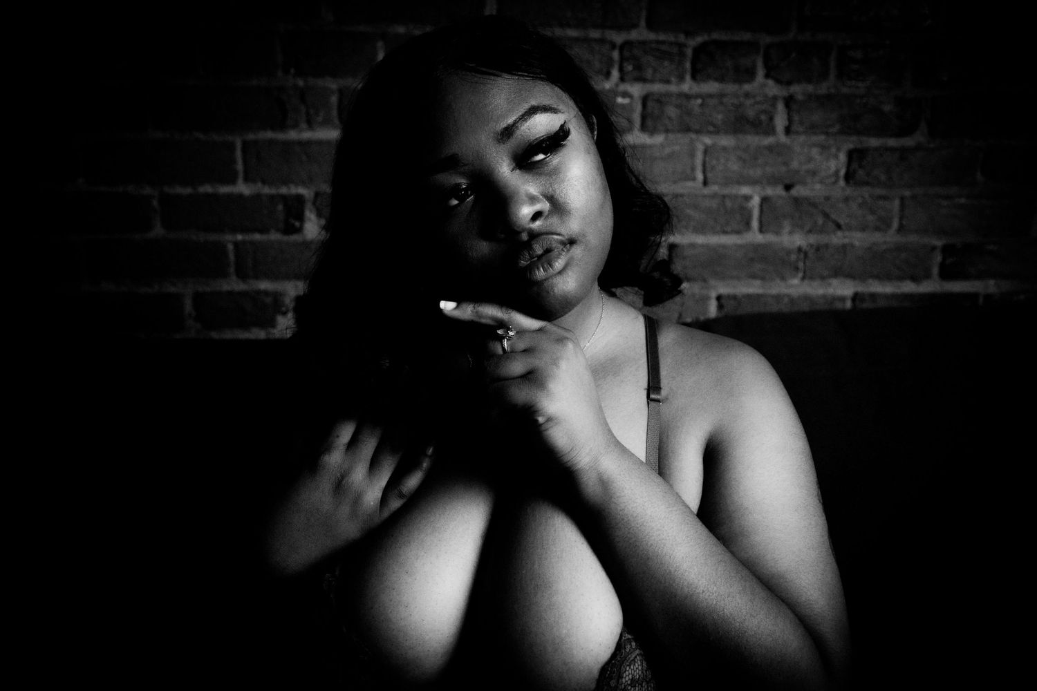 Black and white dramatic portrait of a woman posing against a brick wall in lingerie.