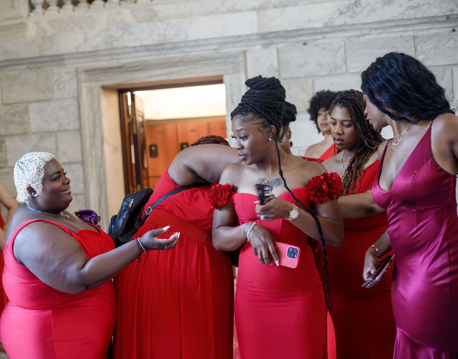 Women in red bridesmaid dresses huddle together examining something in an ornate venue.