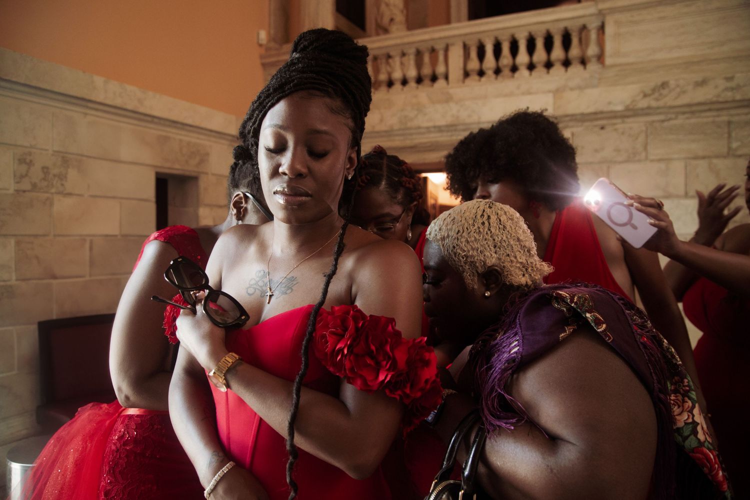 Woman in red off-shoulder dress holds phone while others gather in ornate building interior.