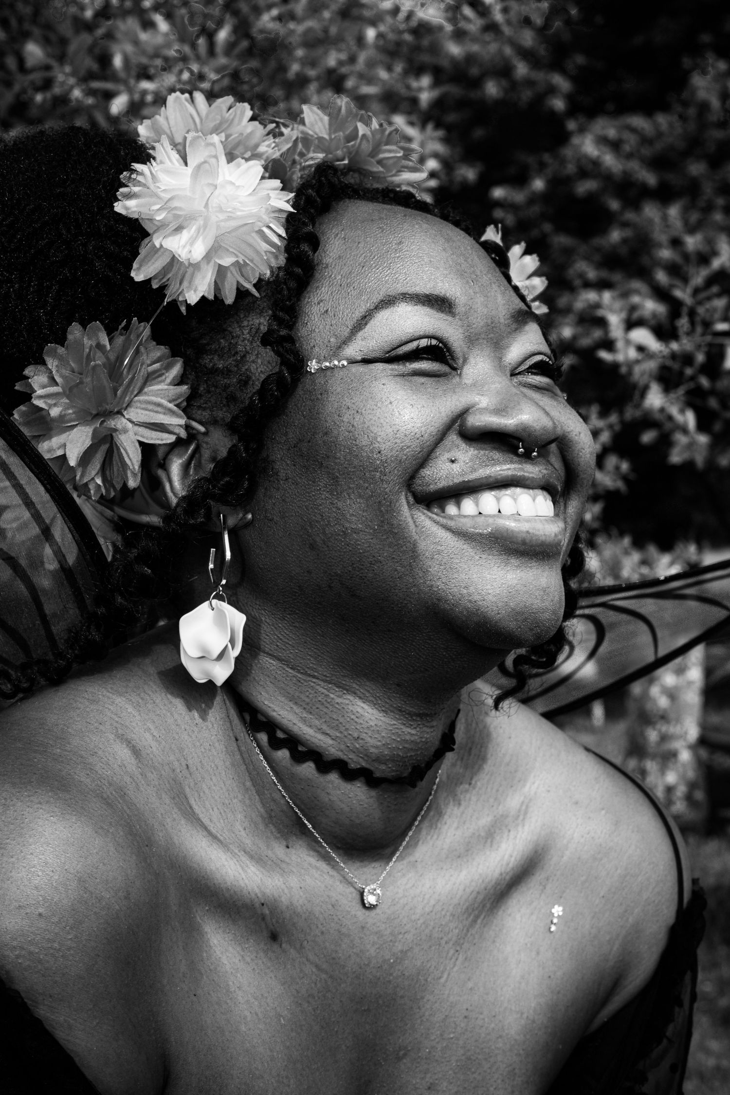 Black and white portrait of smiling woman with flowers in hair wearing earrings and necklace outdoors.