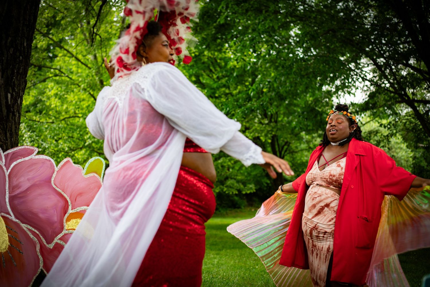 Two women in flowing red and white costumes dance together outdoors surrounded by lush green trees and floral decorations.