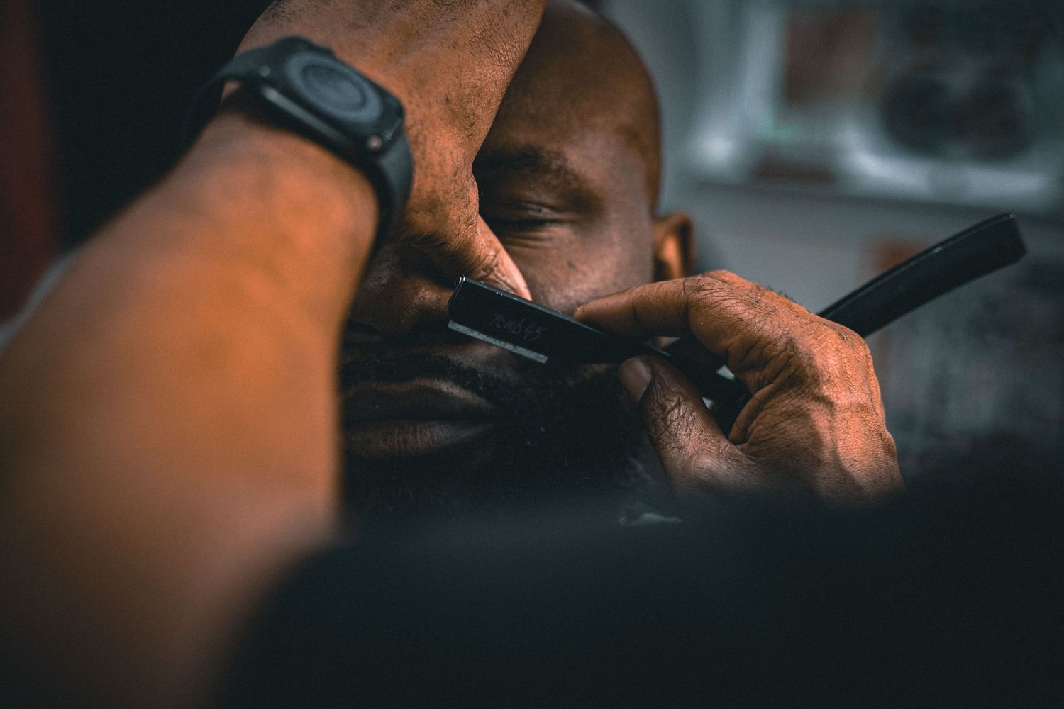 Close-up of barber shaving a mans face with a straight razor in dark lighting.