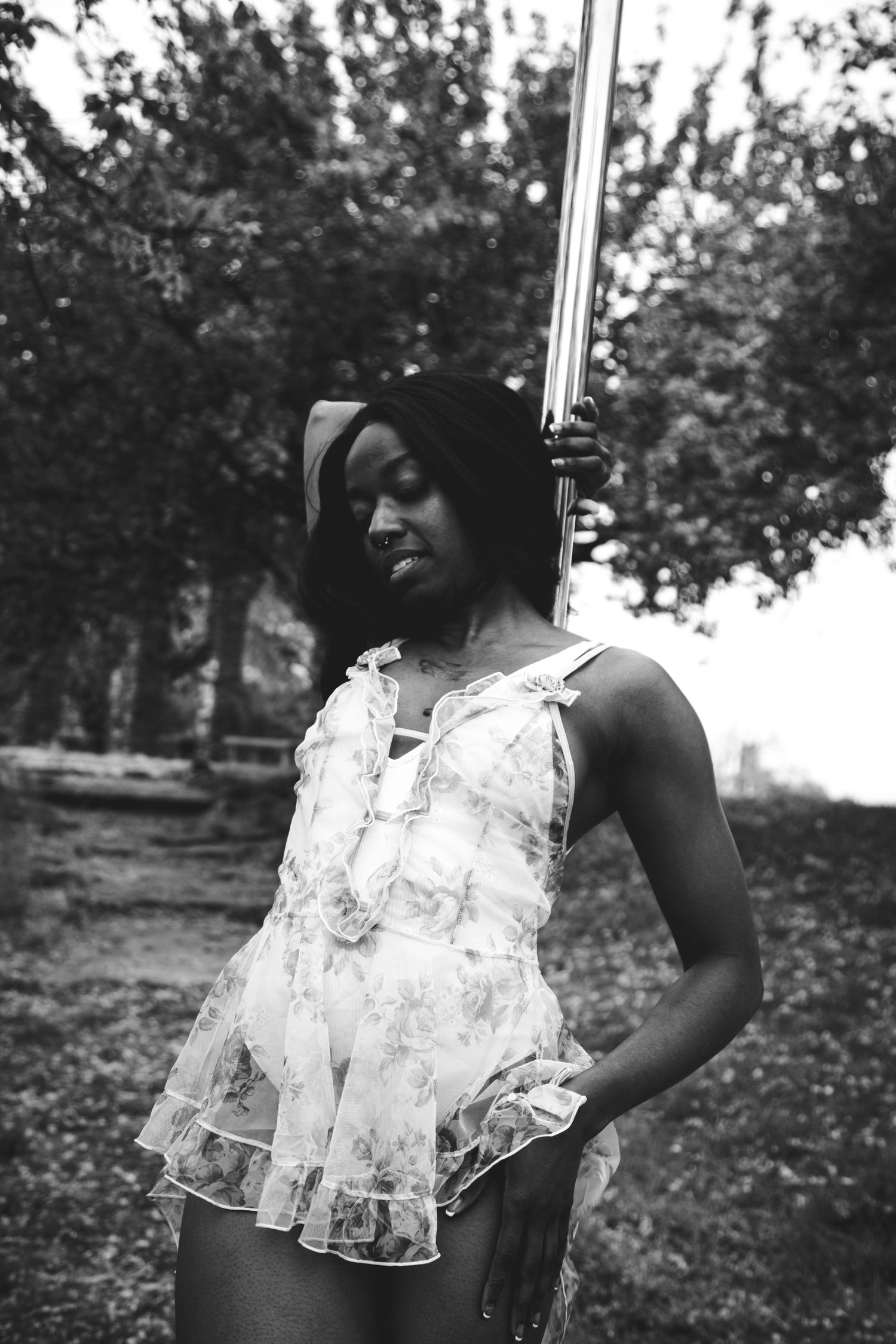 Black and white photo of woman in floral babydoll outfit posing by pole outdoors.