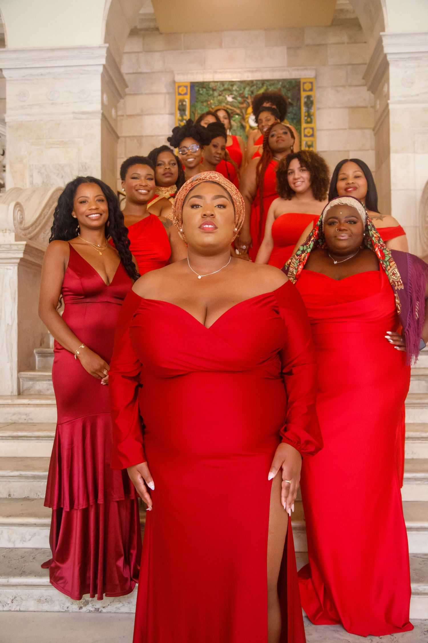 Black women who look like Bridesmaids in red gowns pose on marble stairs with a black woman in gold headwrap leading group.