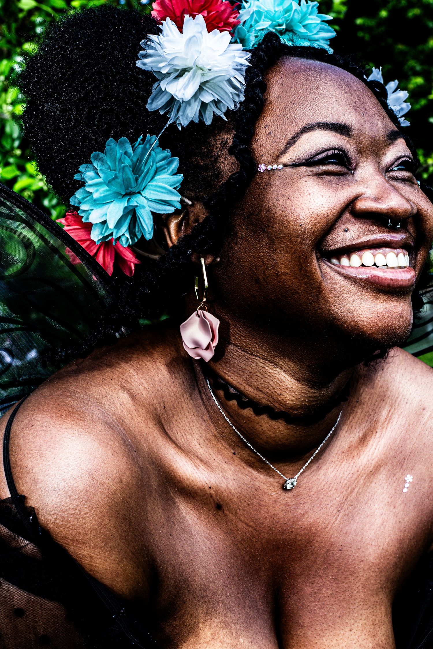 Joyful woman laughing with colorful flowers in natural hair wearing necklace at outdoor festival.