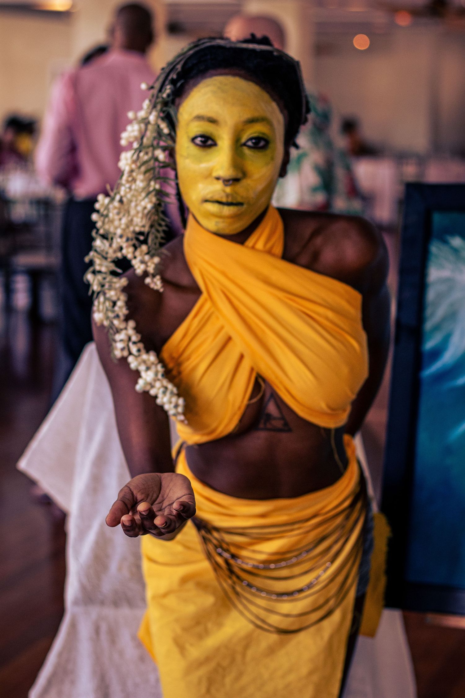 Model with yellow face paint wearing orange draped costume and beaded chains extending open hand at indoor event.