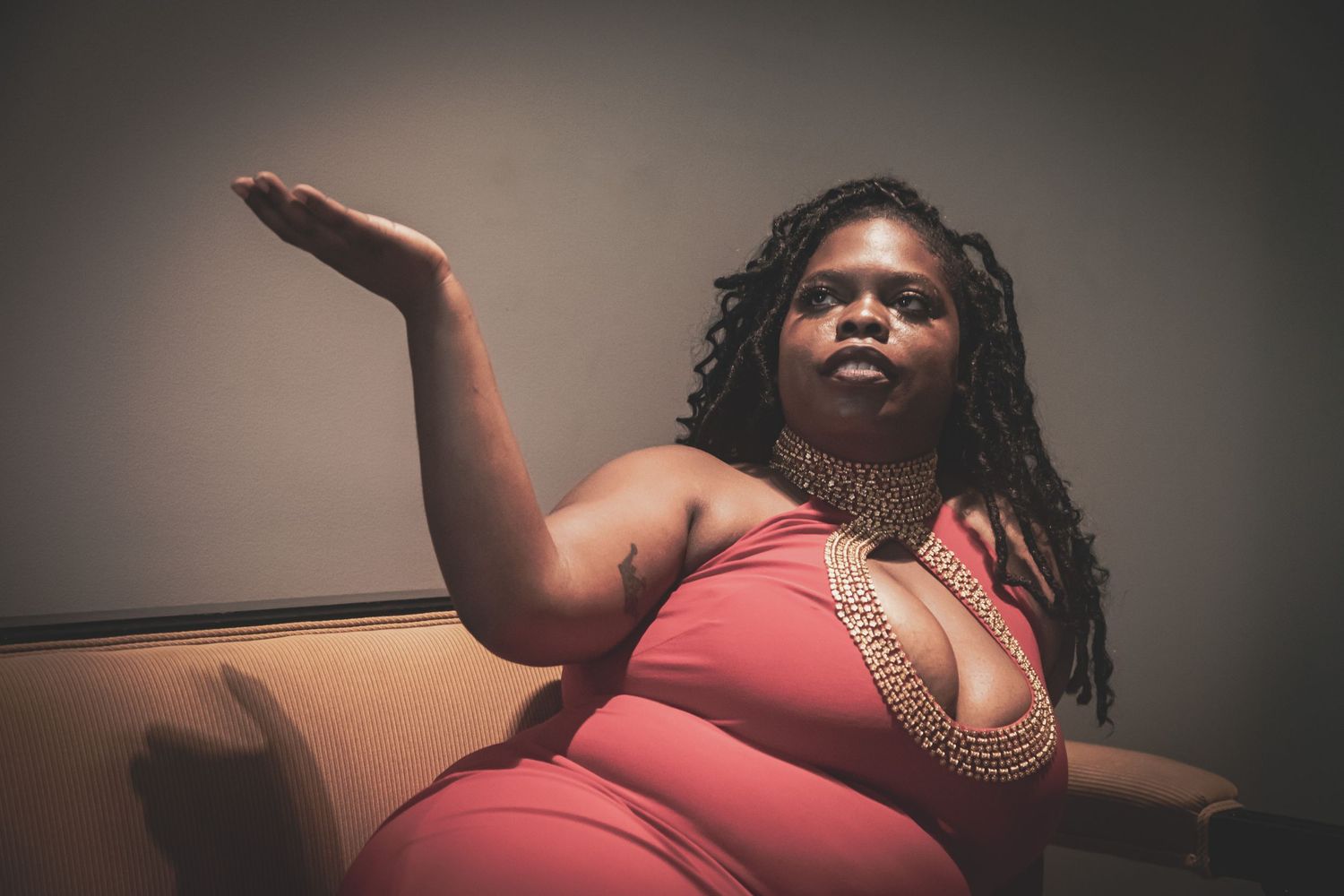 A plus-size Black woman in a red outfit with beaded necklace poses on a sofa, arm extended.