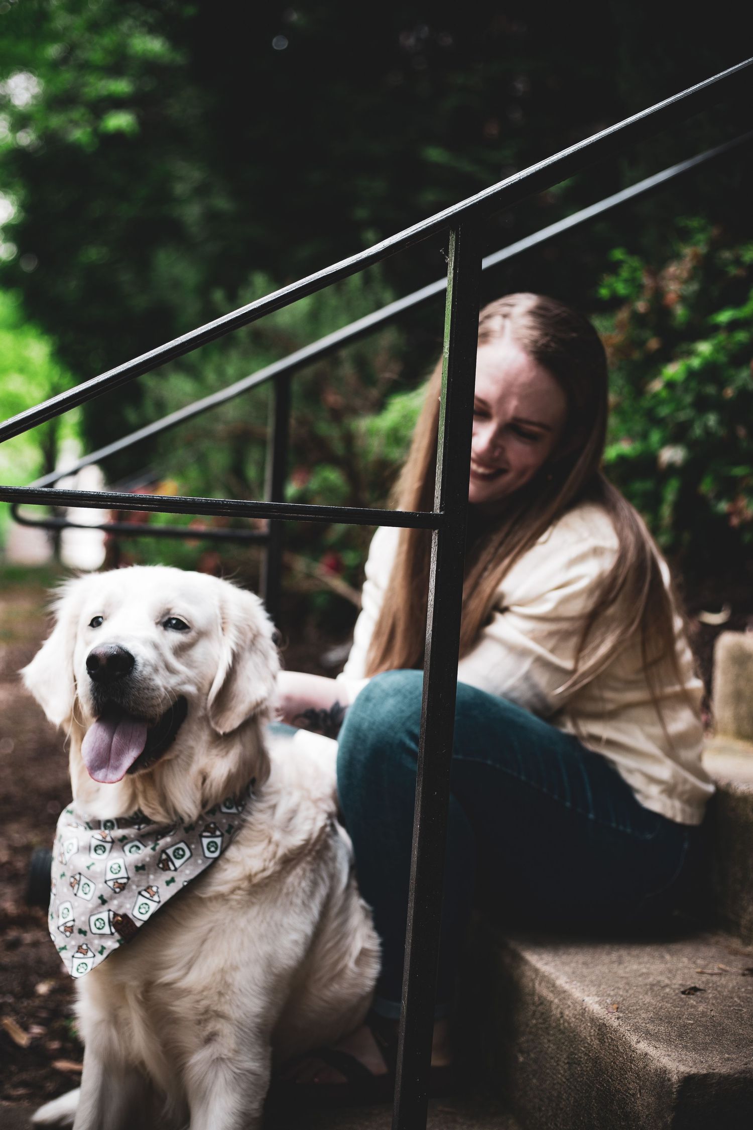 A smiling woman sits on stone steps petting a fluffy golden retriever wearing a bandana outdoors.