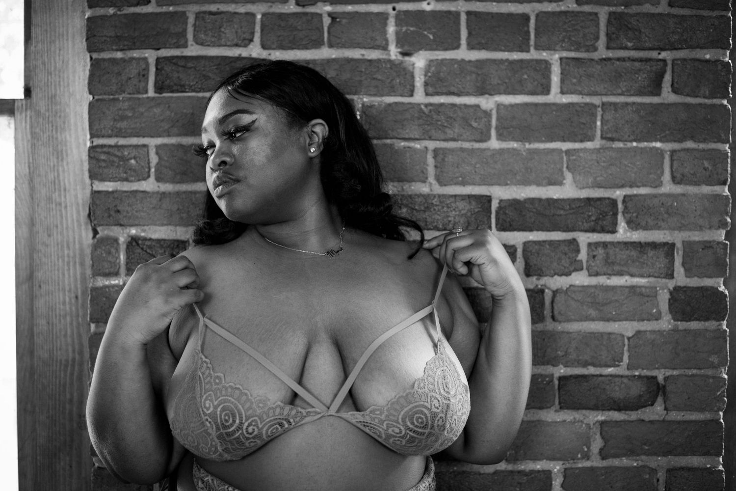 Black and white photo of a plus-size woman in a strappy lace bra posing against a brick wall.