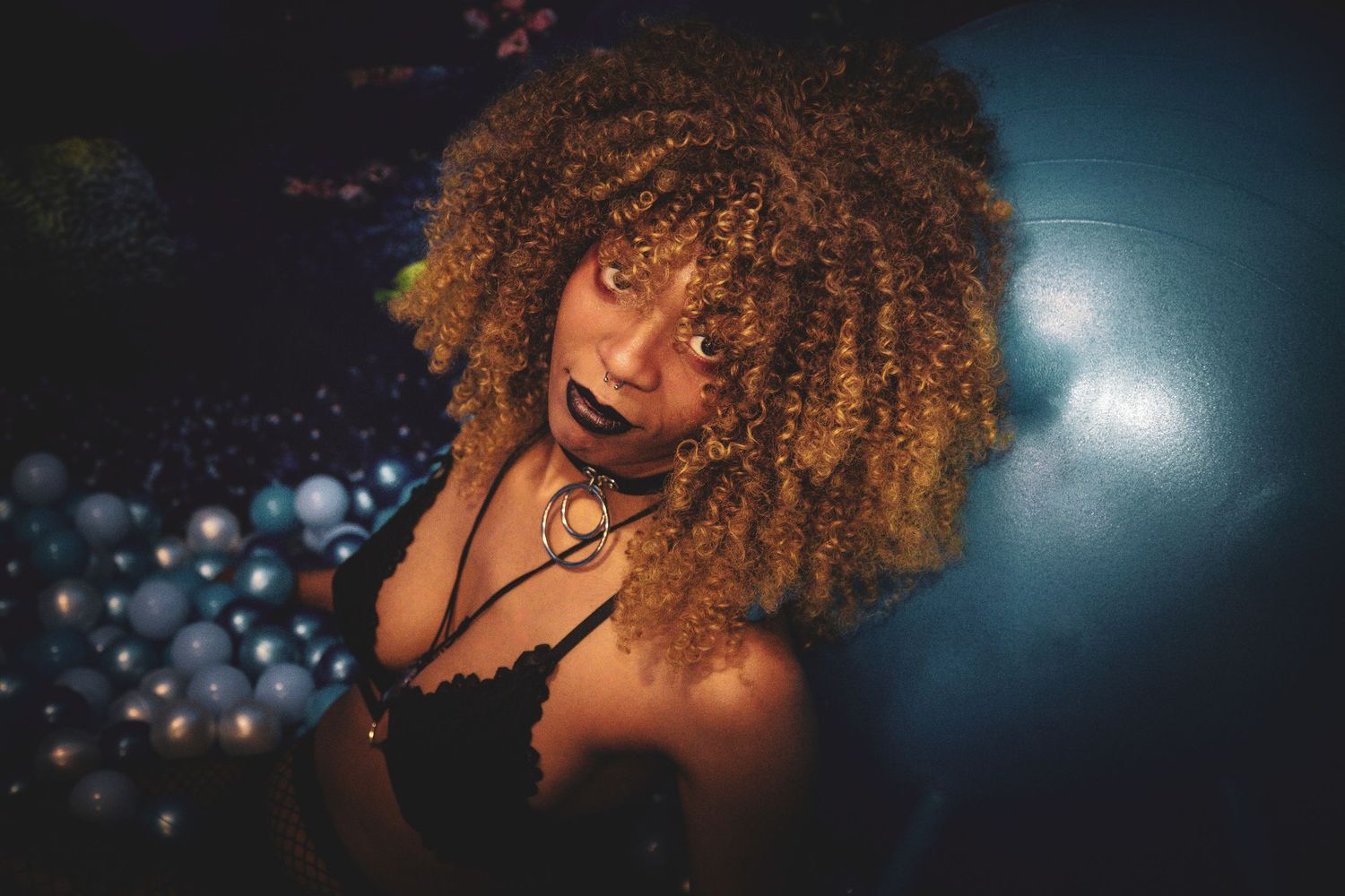 Woman with voluminous curly auburn hair and dark lipstick posing in dramatic blue lighting.