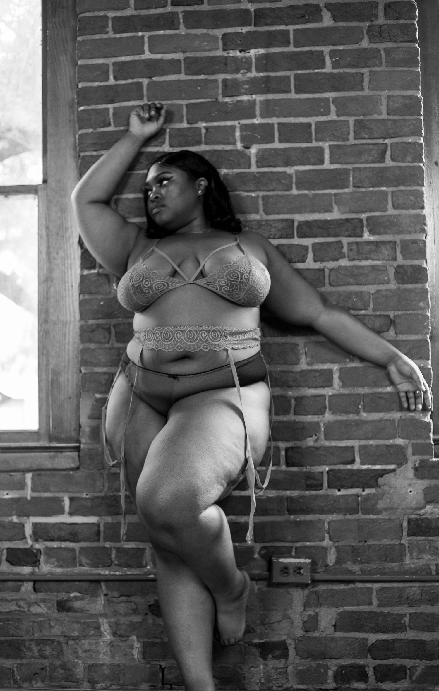Black and white portrait of a plus-size woman in lace lingerie posing against a brick wall.