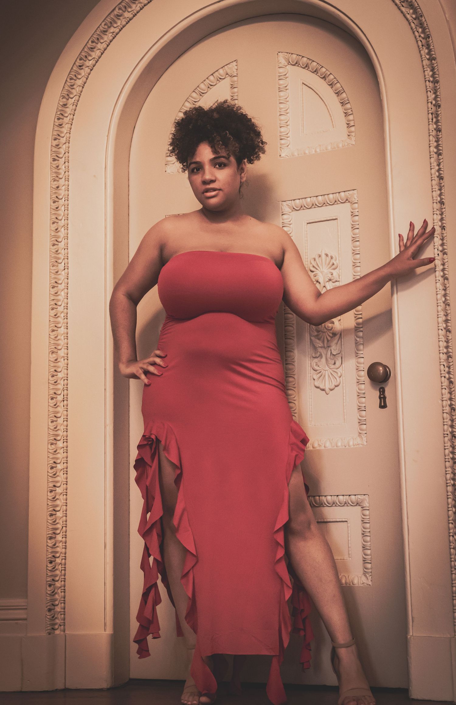 Curly-haired woman in a red ruffled strapless dress poses elegantly beside an ornate arched white door.