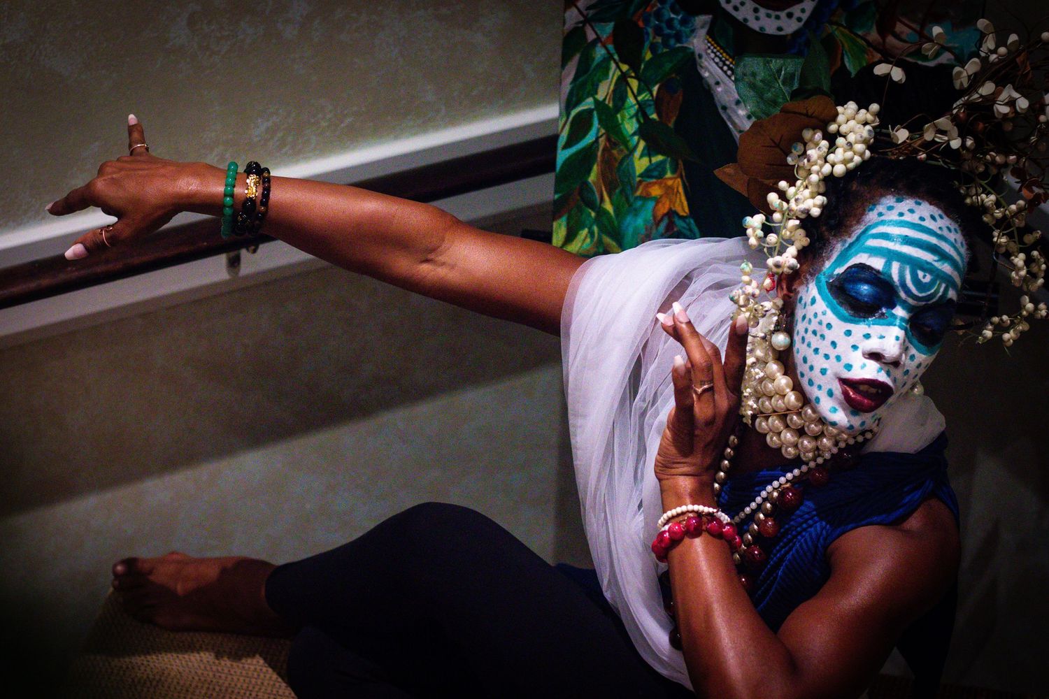 Woman in blue dotted face mask, floral crown, and pearl necklaces posing dramatically on stairs.