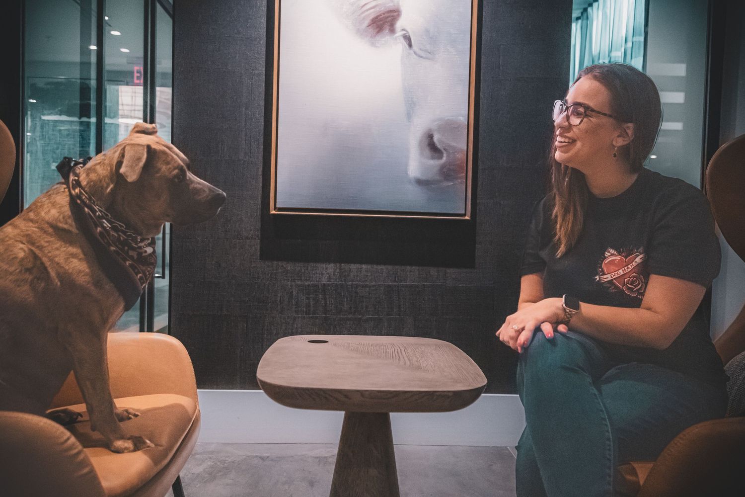 Smiling woman in glasses sitting across from a brindle, pitbull-mix dog at a small table indoors.