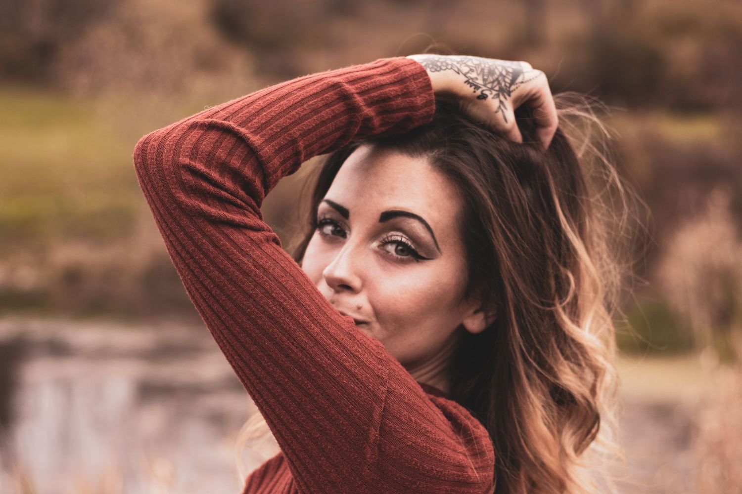 Young woman with tattoos in rust sweater poses outdoors with hands in windswept hair.