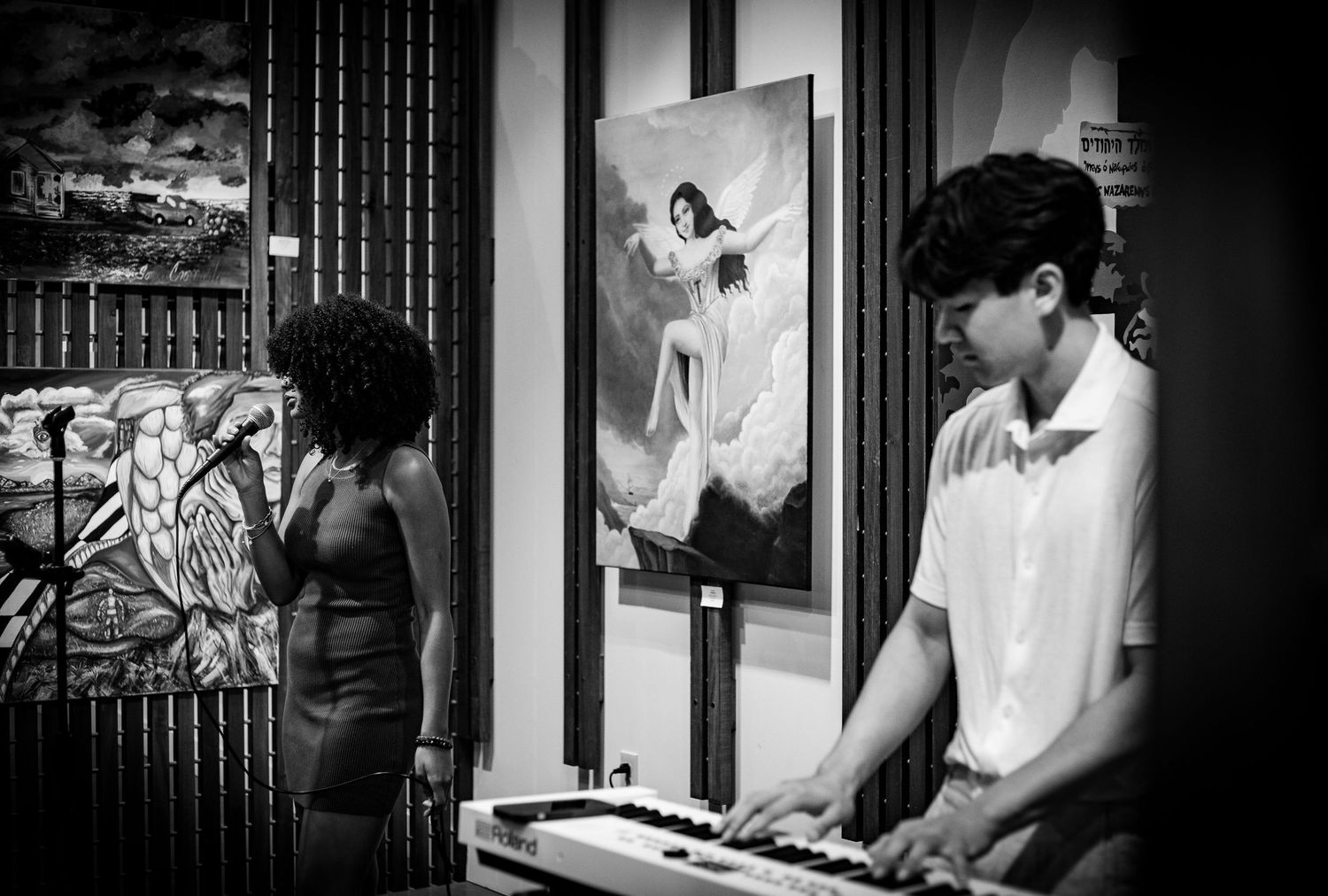 Black and white photo of a young man playing keyboard at an art gallery event with paintings on the wall.