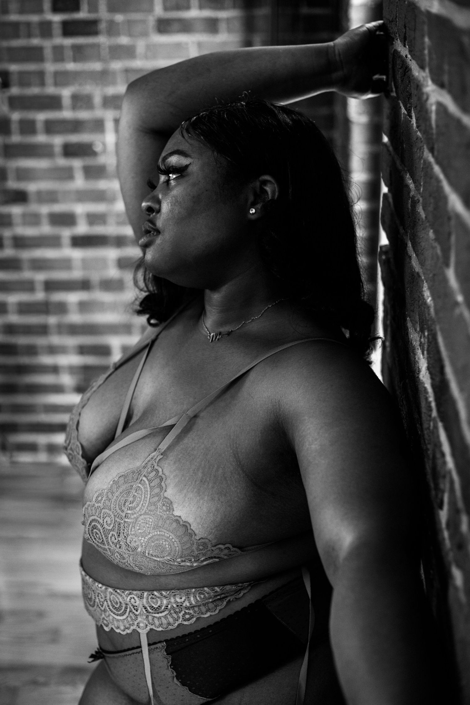 A Black plus-size woman leans against a brick wall in a lace bralette, arm raised, in a black and white portrait.
