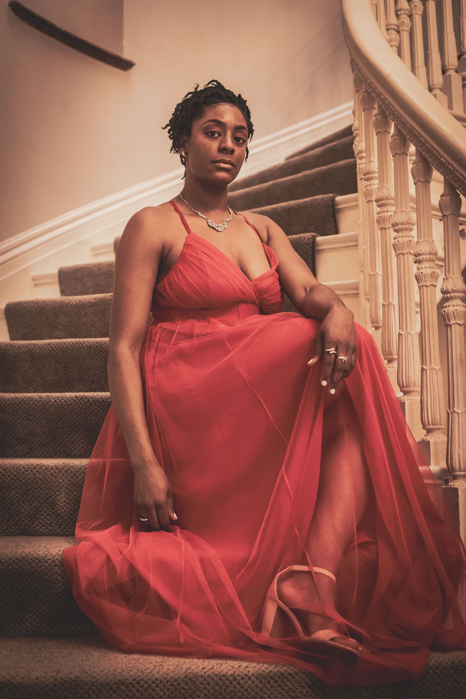 Elegant Black woman in a flowing coral tulle gown sits gracefully on carpeted stairs with white banisters.