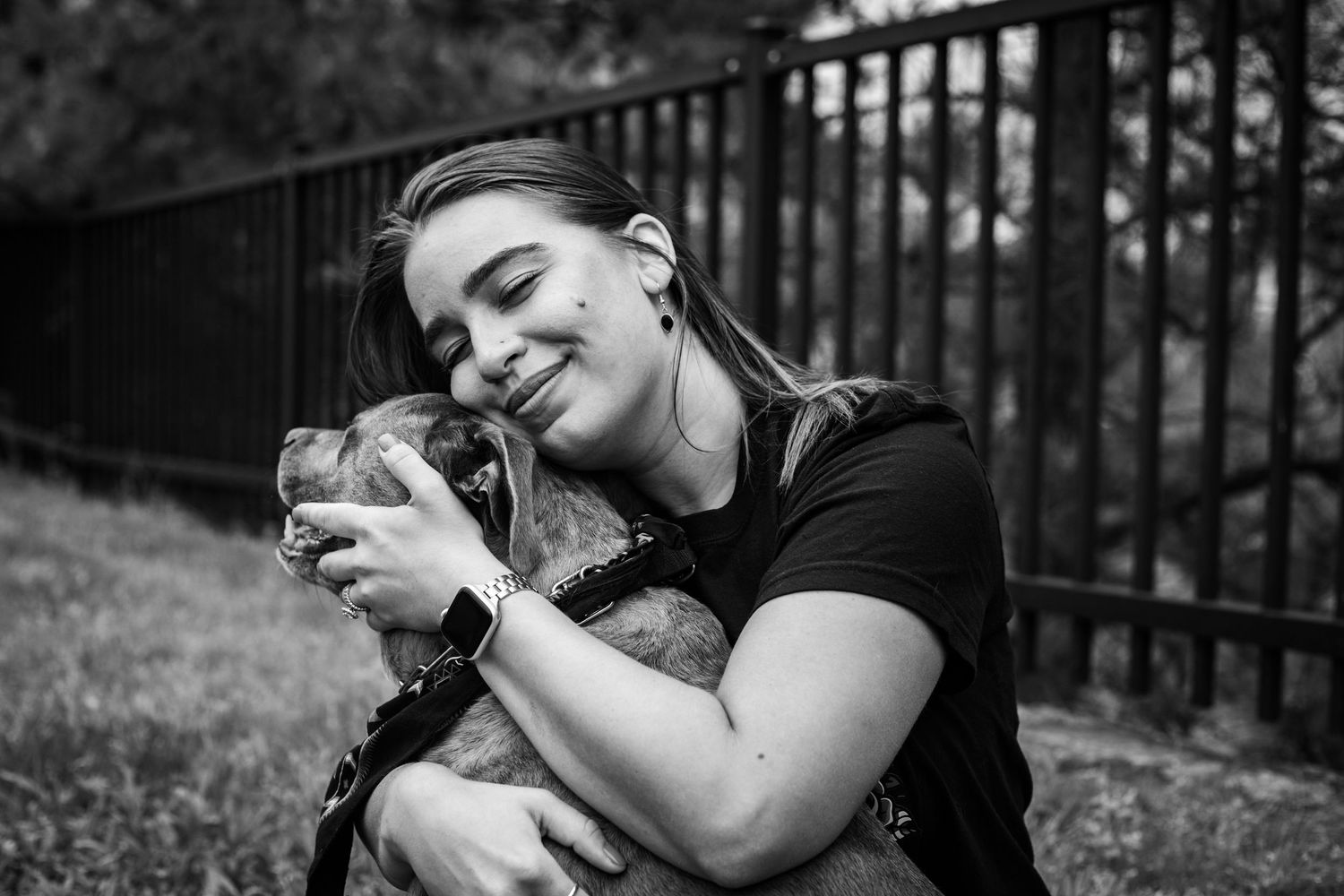 Black and white photo of smiling young woman joyfully hugging her brown dog outdoors.