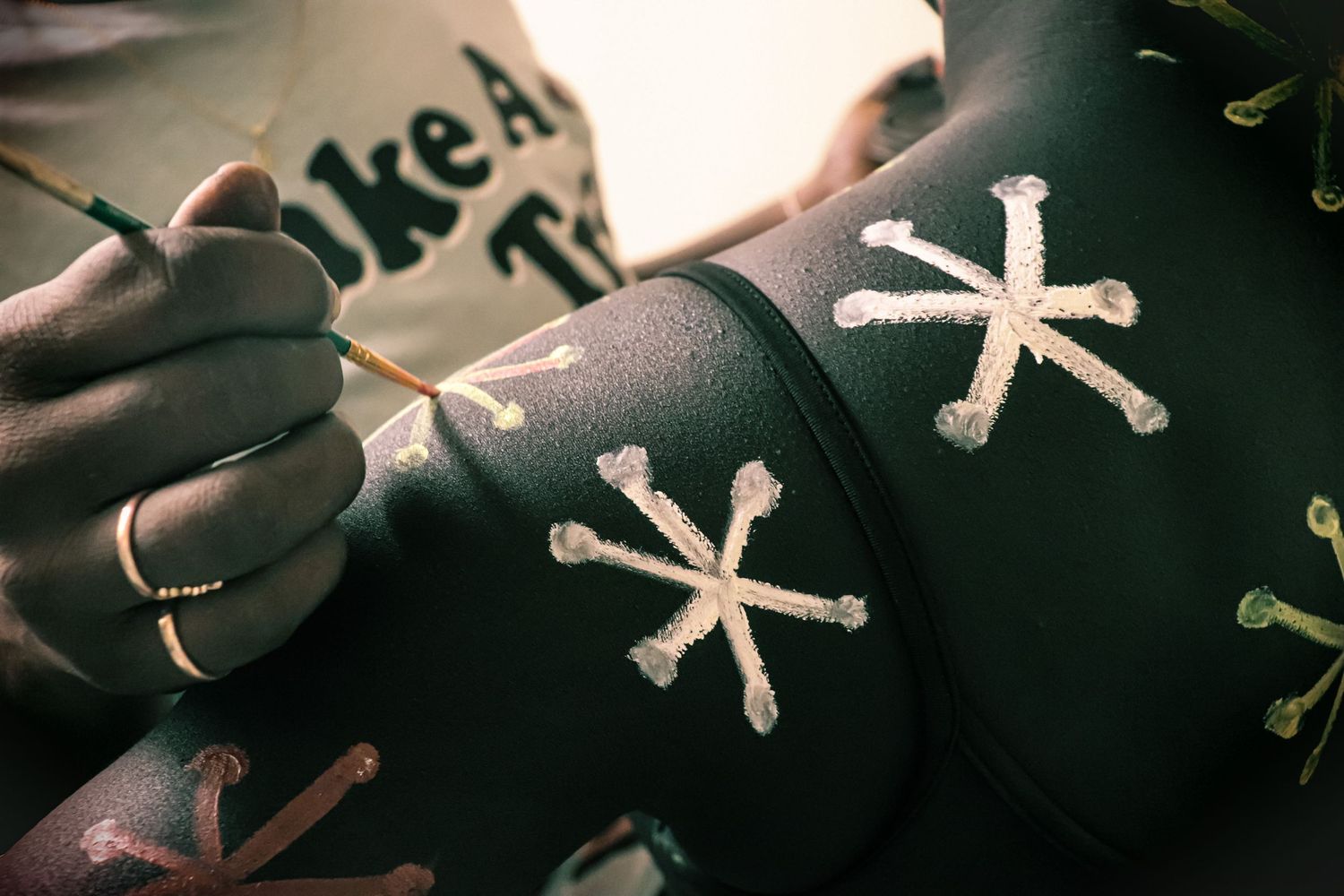Artist painting white asterisk star designs onto dark body-painted skin using a fine brush.