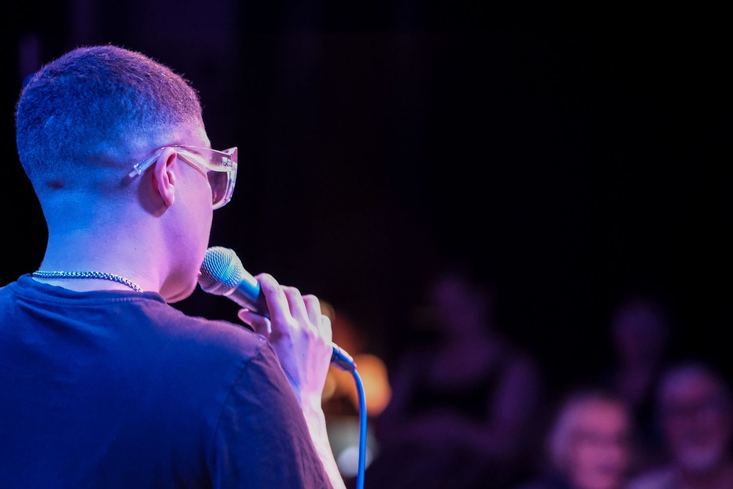 Rear view of a bald man wearing glasses speaking into a microphone on a lit stage.
